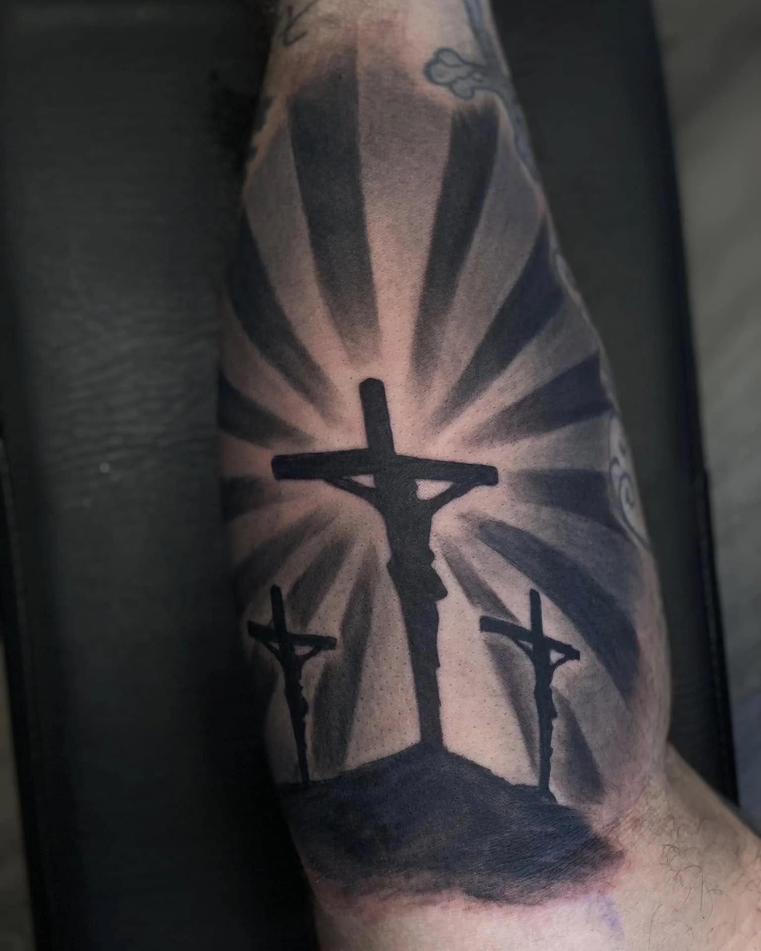 A tattoo on a person's arm featuring a large cross with rays emanating from it, with additional elements including a smaller cross and a head of a dog or wolf above.