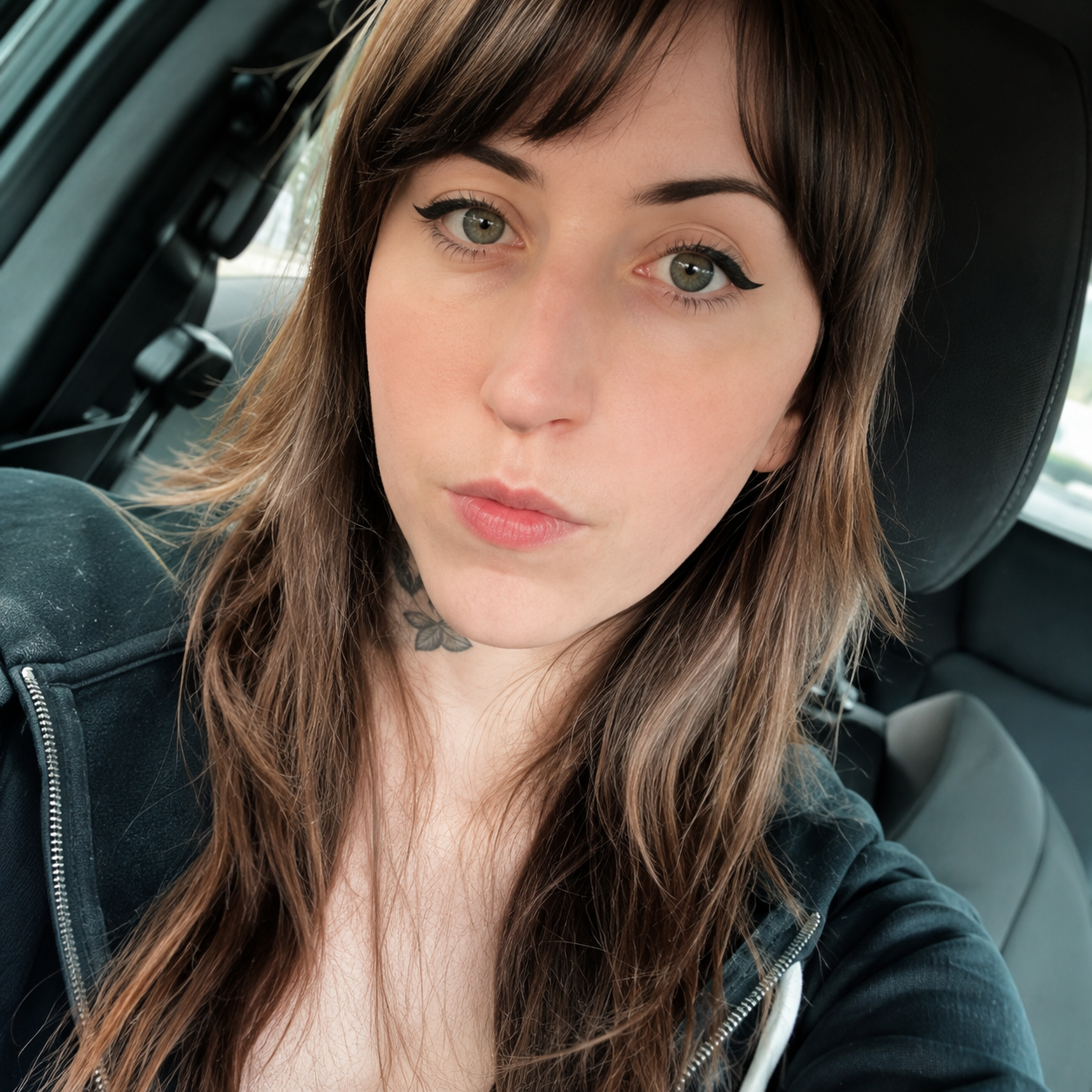 Close-up of a young woman with brown hair and green eyes sitting in a car, wearing a black jacket and makeup with winged eyeliner, looking into the camera.