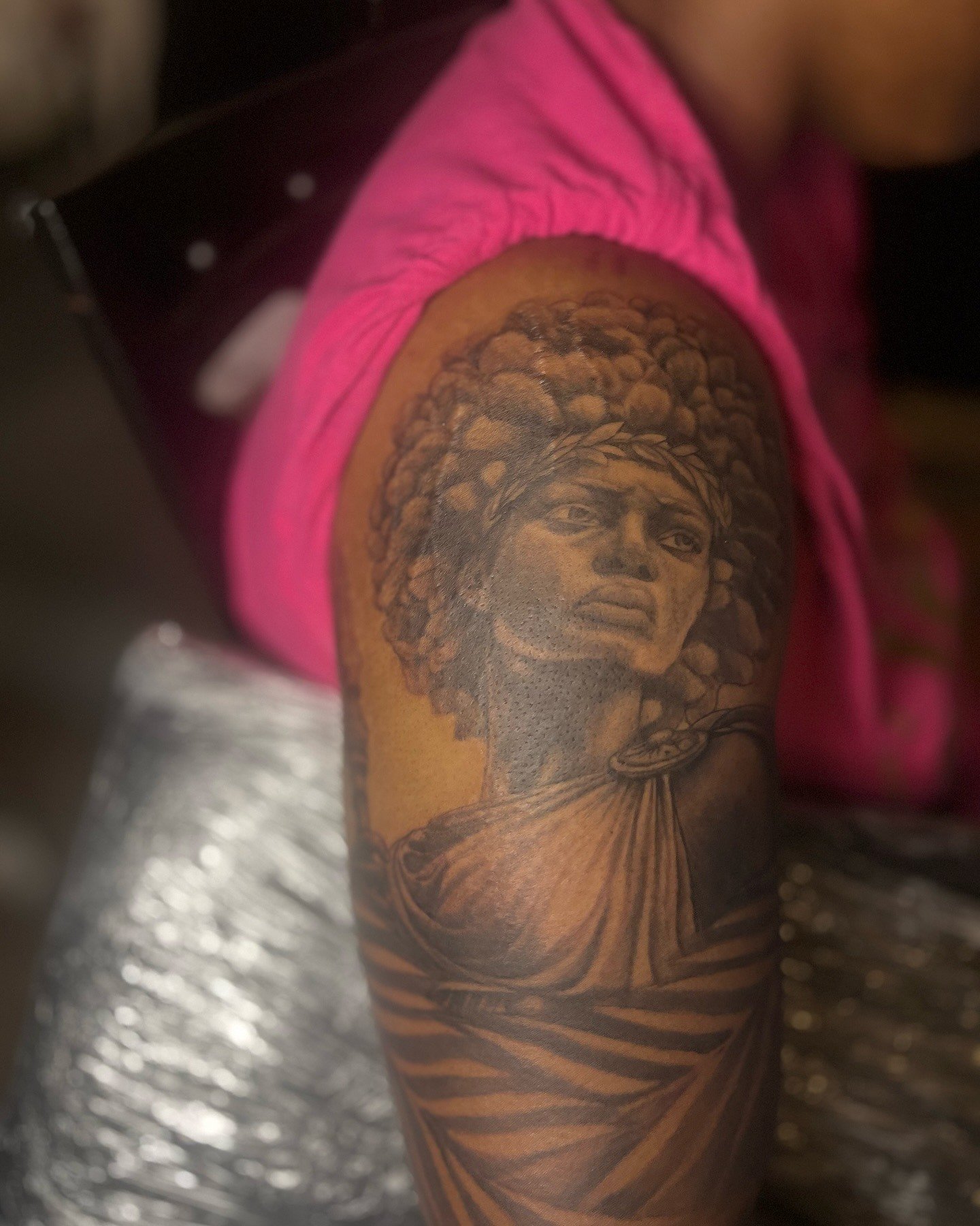 Tattoo of a woman with an intricate feathered headdress on a person's arm, with a pink shirt in the background.