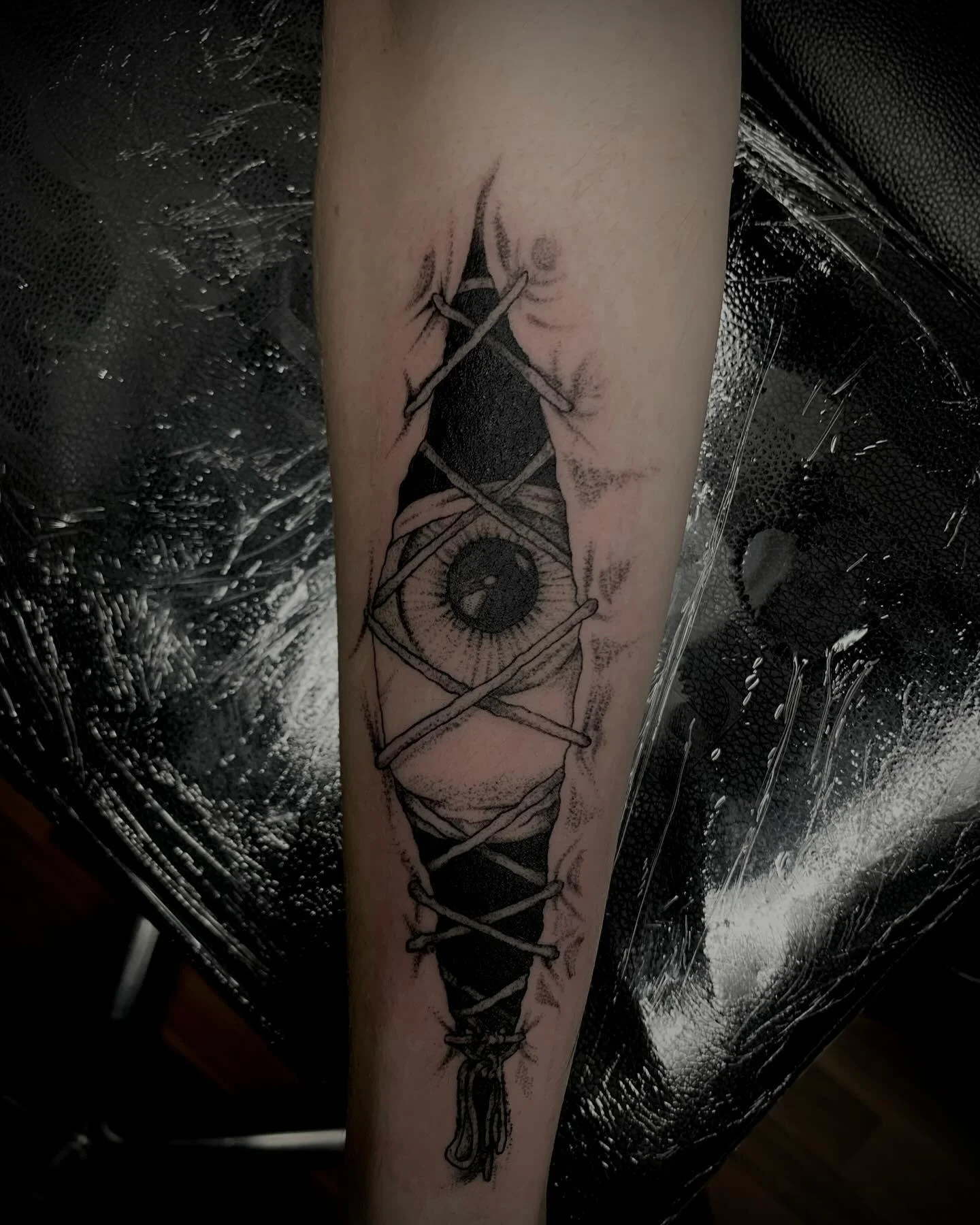 Black ink tattoo of a camera with a lollipop in the center, wrapped with laces, on person's forearm.