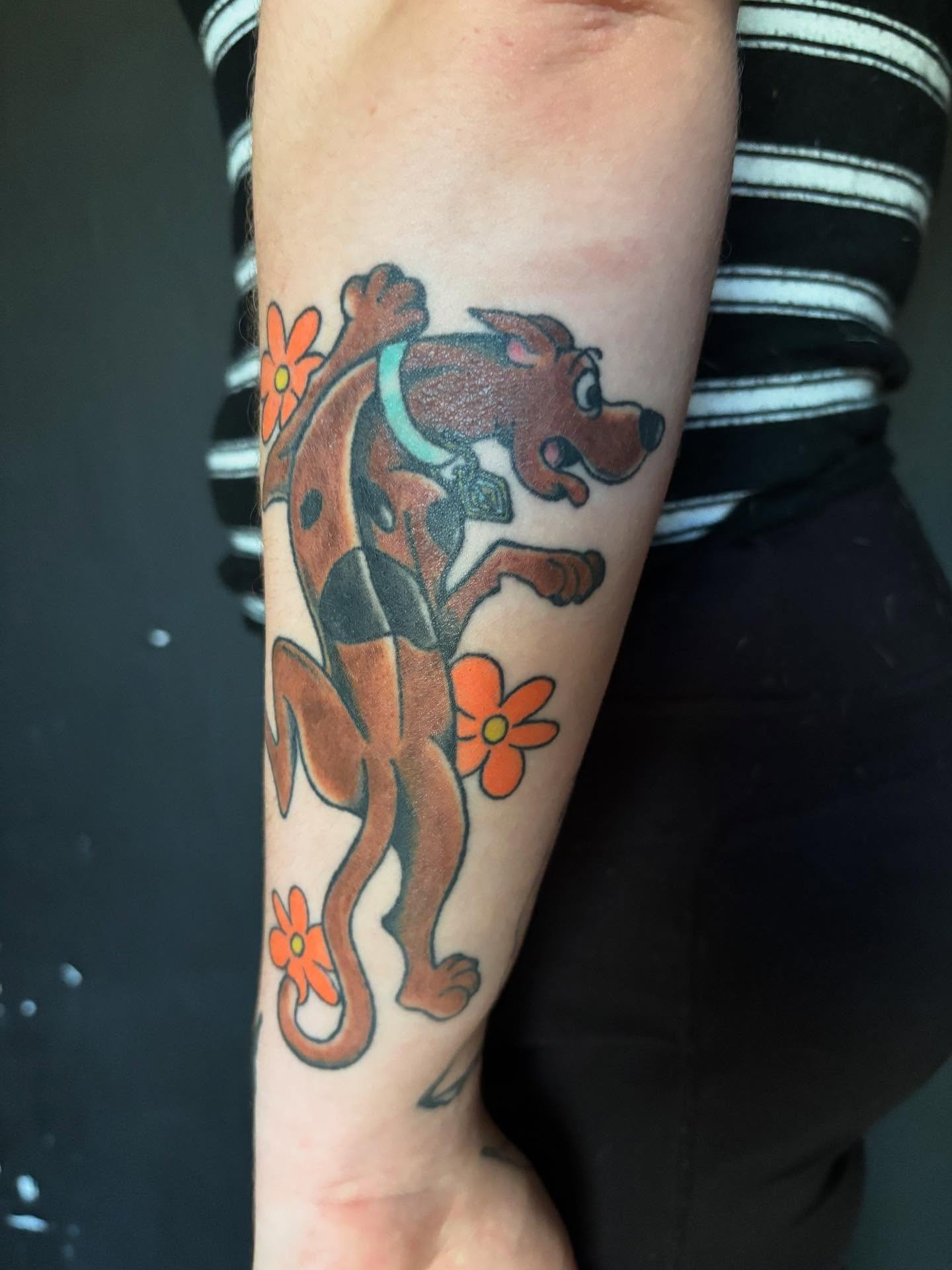 Tattoo of Scooby-Doo with two orange flowers on the person's forearm.