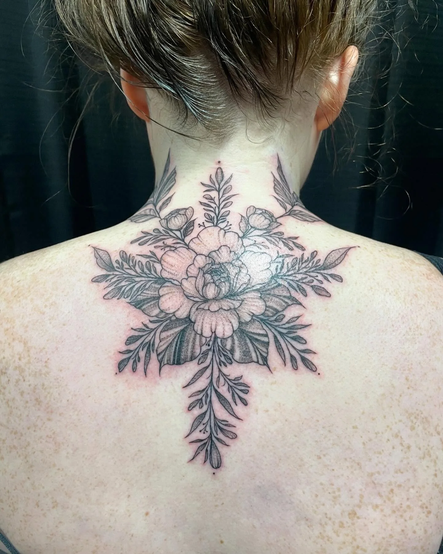 A person's back with a black and gray floral tattoo extending from the neck downward, featuring large flowers and leaves.