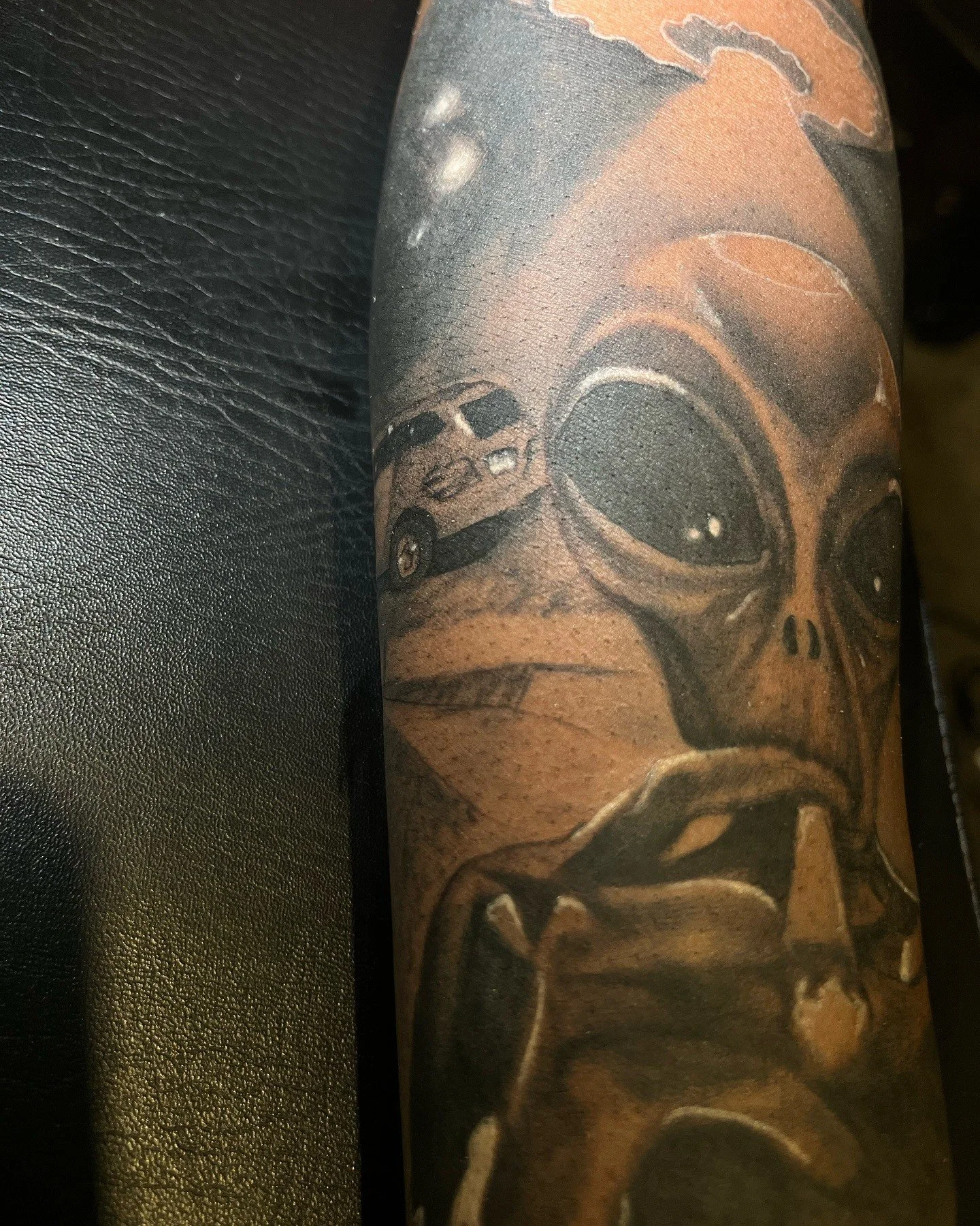 Close-up of a tattoo of a skull wearing sunglasses on a person's arm, with a background of a black leather surface.