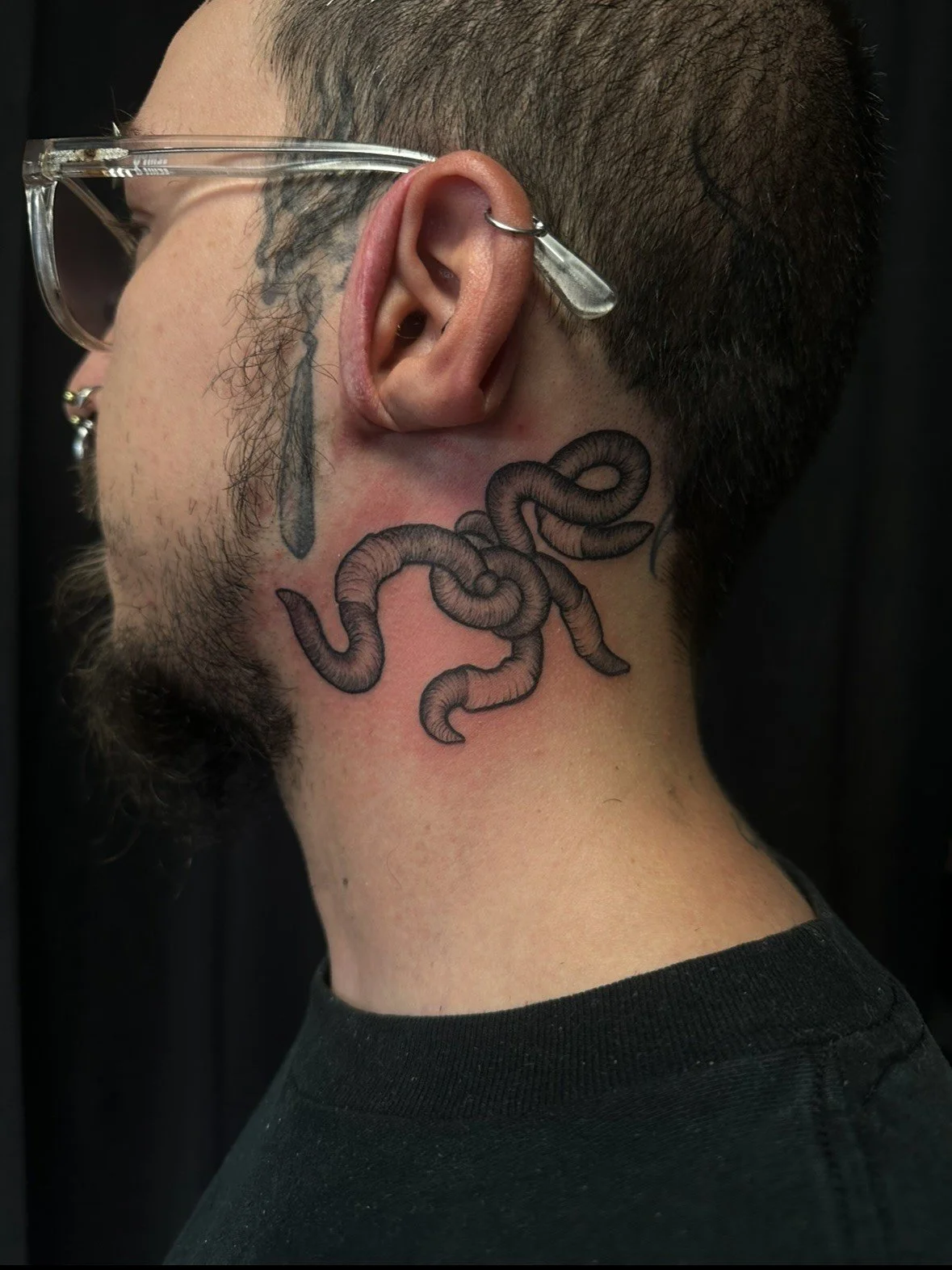 Close-up side of a person's neck with a black tattoo of a coiled worm and additional smaller details, wearing clear glasses and earrings, with short dark hair and facial hair.