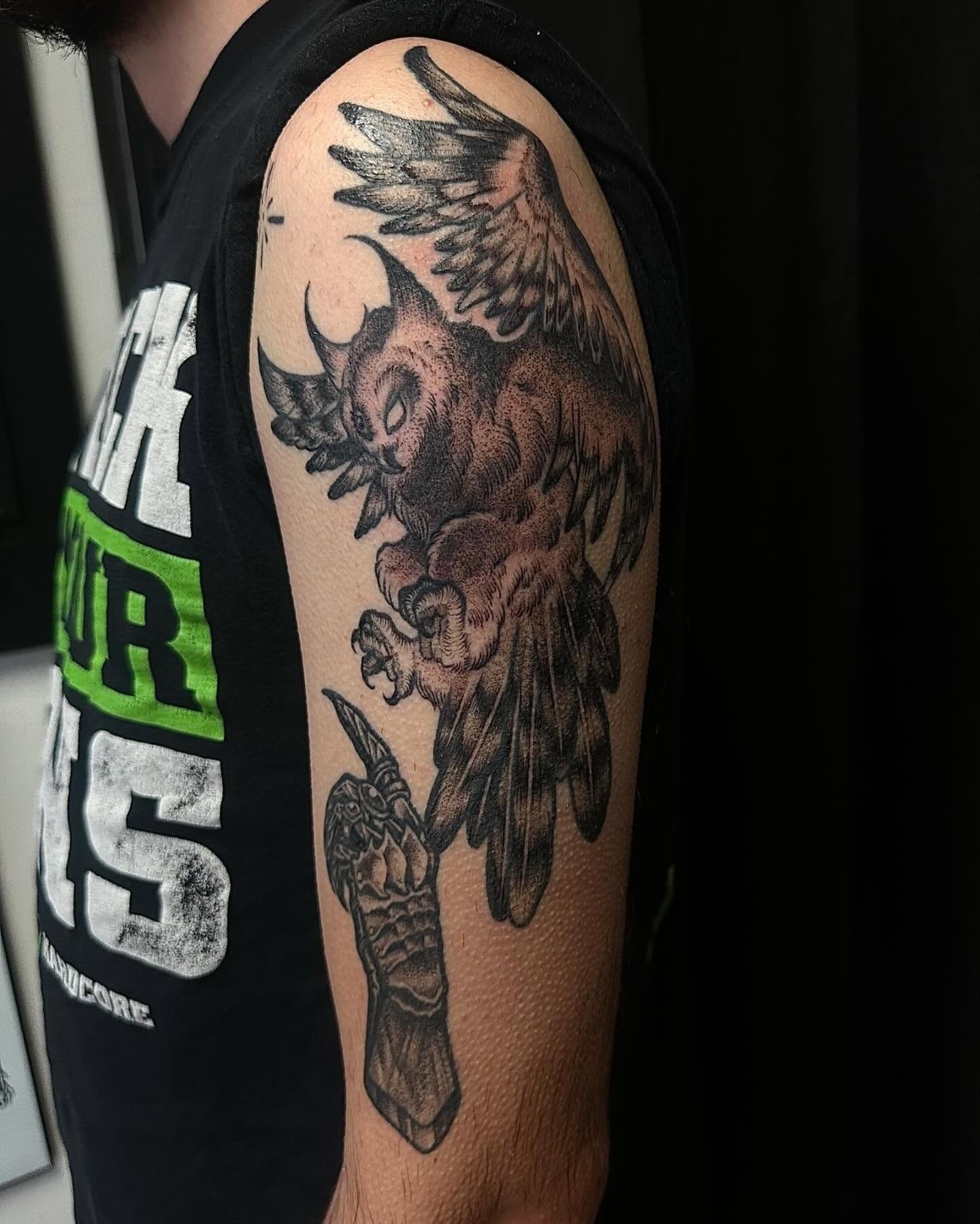 Tattoo of a mythical creature with the head of a wolf, wings, and a sword in its paw on a person's upper arm.