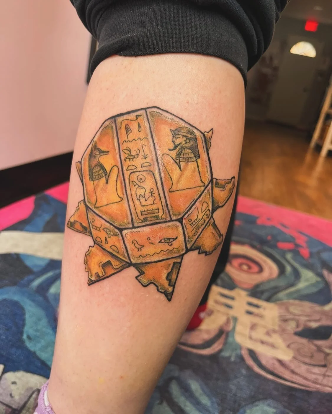Tattoo of a colorful ancient Egyptian scarab with hieroglyphs and animal figures on a person's thigh.