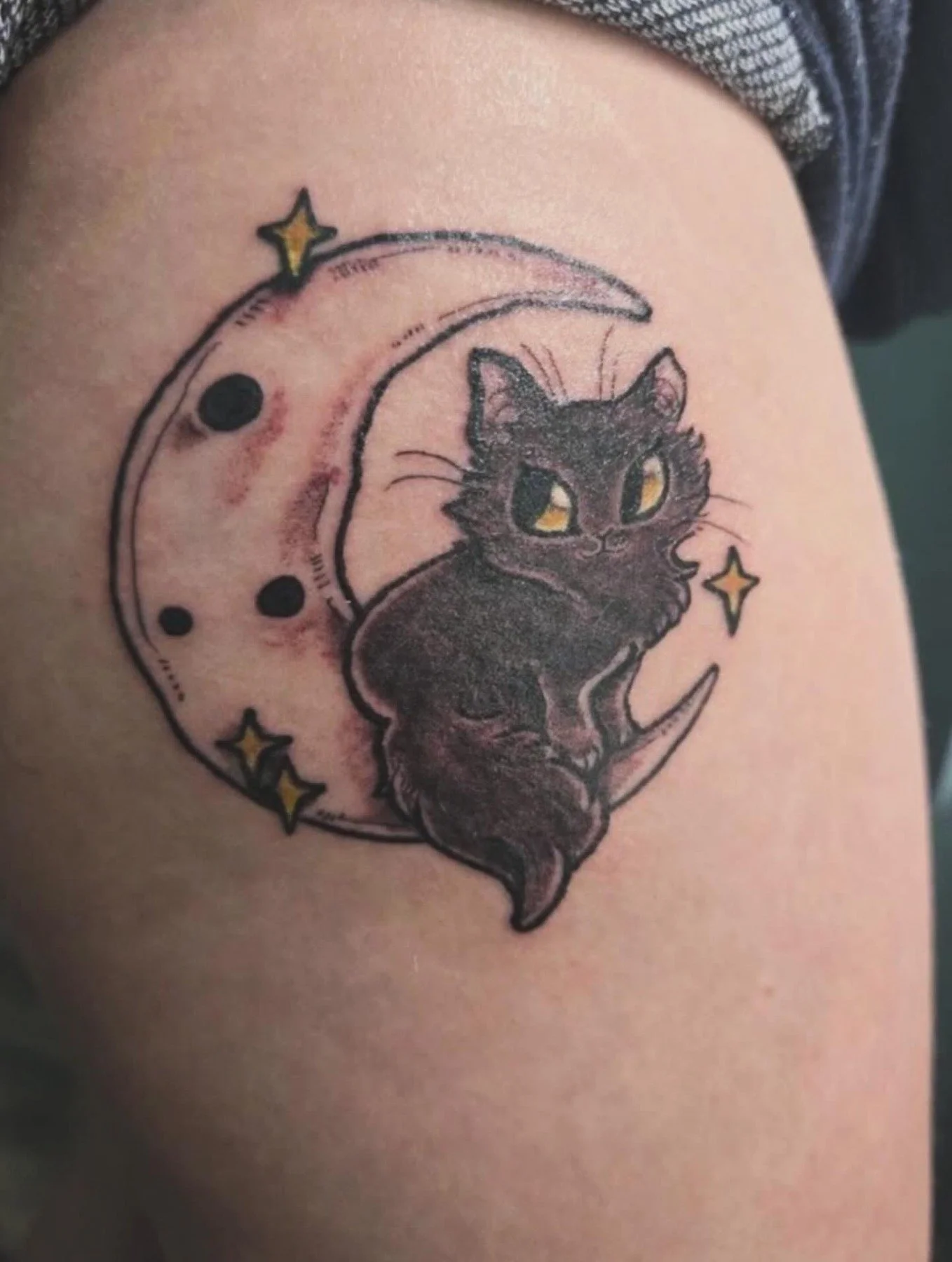 Tattoo of a black cat sitting on a crescent moon with three small stars around it.