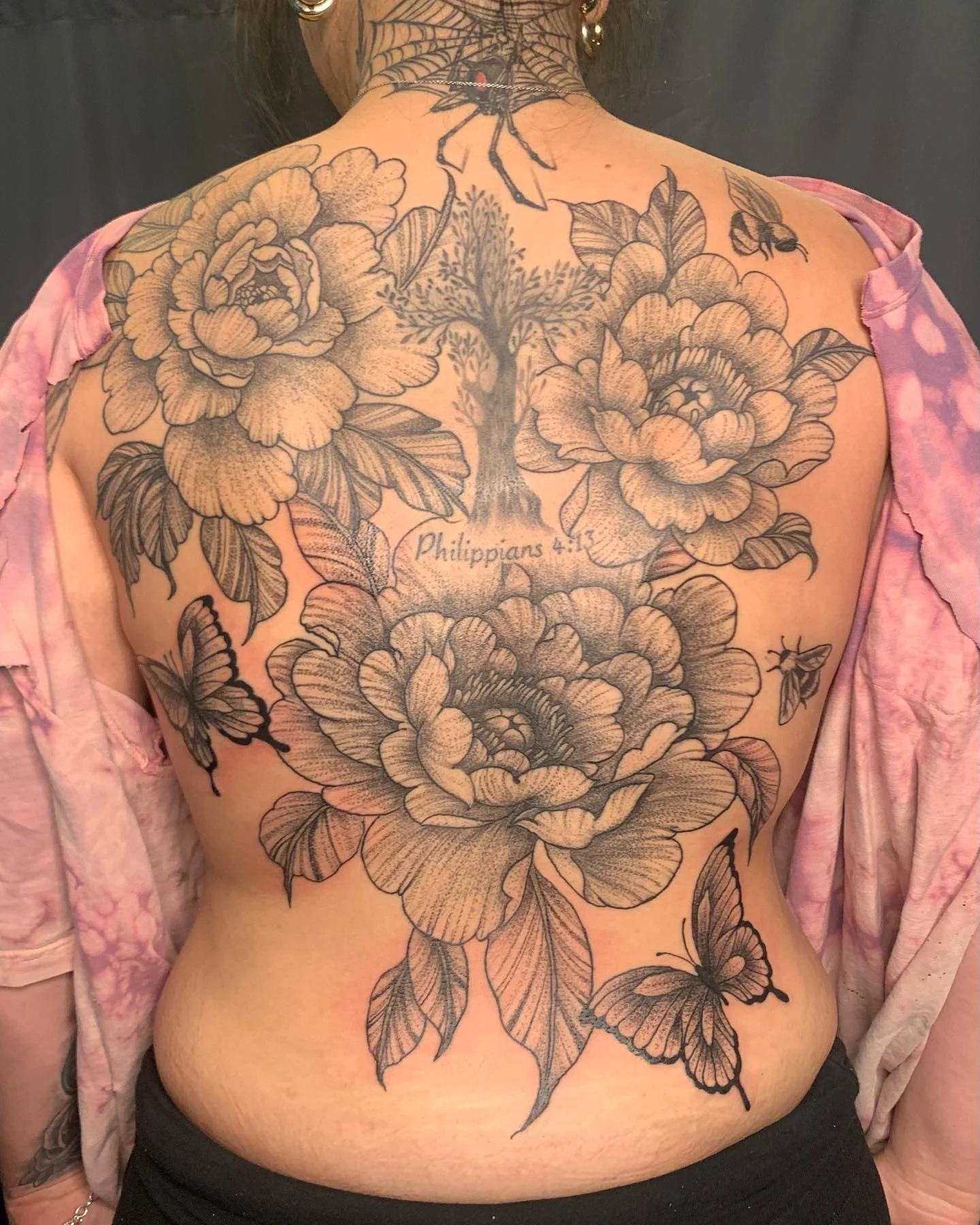 Back view of a person with extensive black and gray floral tattoos covering the upper back, shoulders, and neck, including large peony flowers, butterflies, and a spider with a web. The person is wearing a pink and white patterned garment off the sho