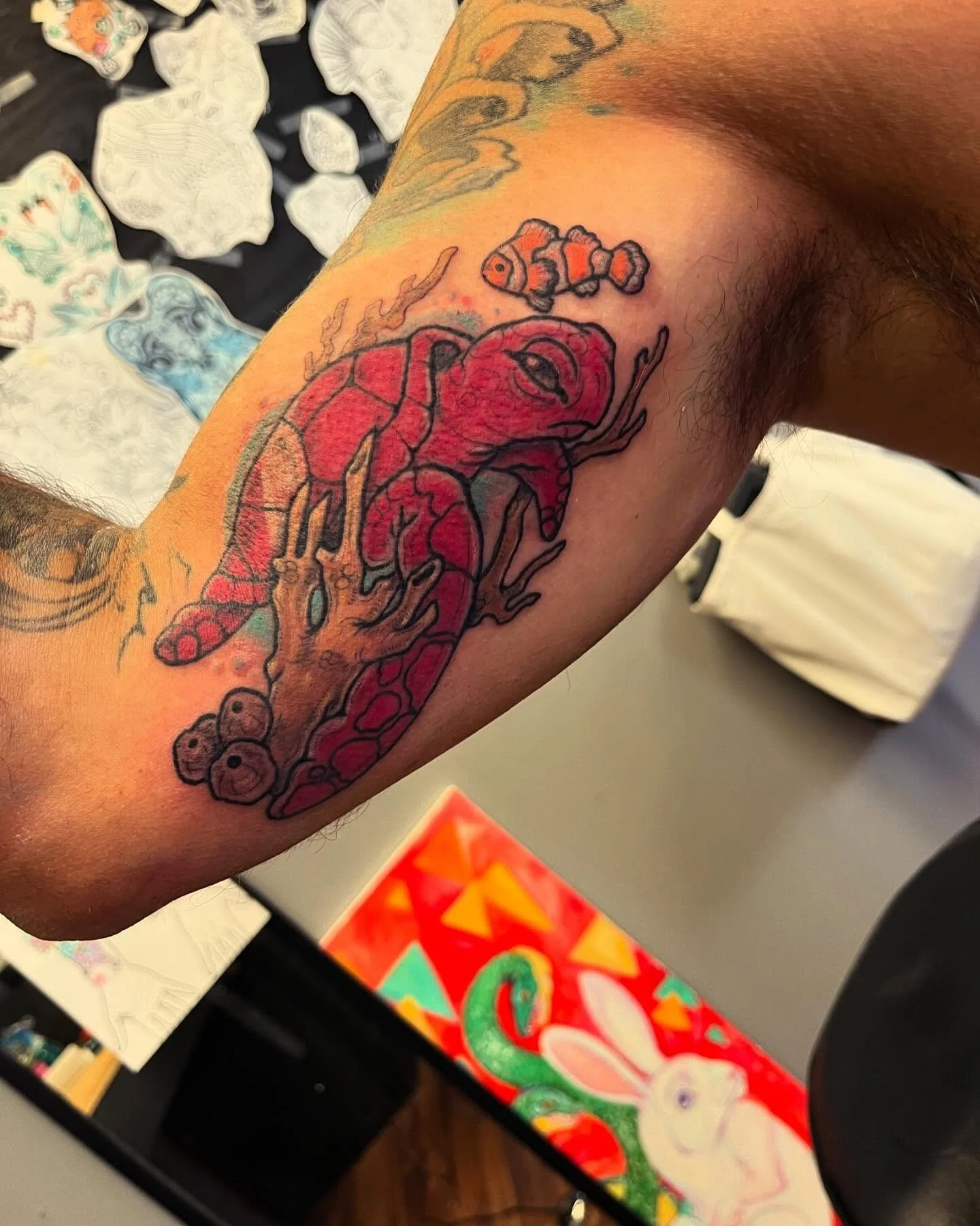 A tattoo of a red crab with detailed black outlines on a person's forearm. A small clownfish is above the crab. Tattoos of various other designs are visible in the background.