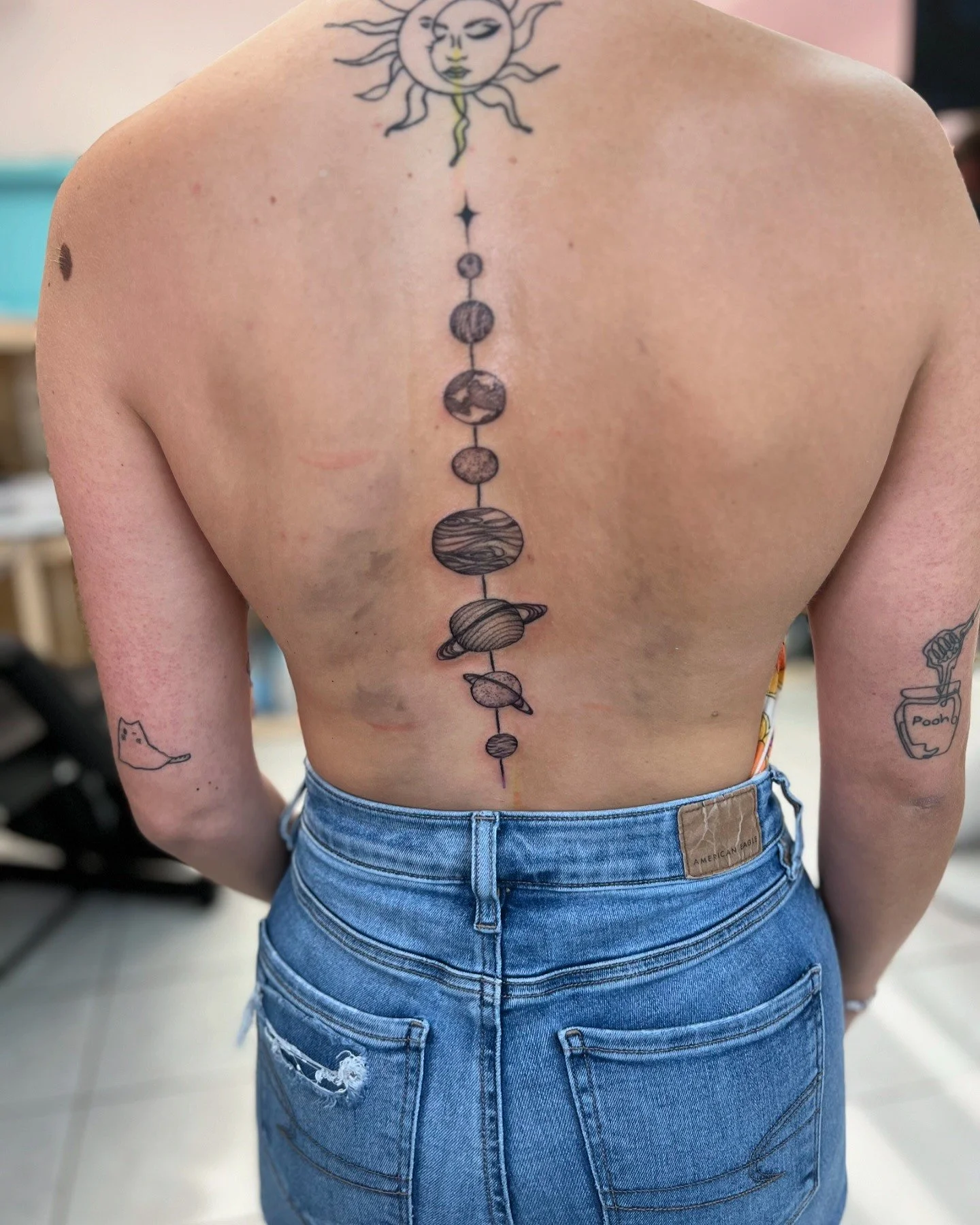 A person's back with a vertical tattoo of the solar system, surrounded by other tattoos including a sun and moon face at the top, a bird on the left arm, and a plant with the word "Pooh" on the right arm, wearing blue jeans.