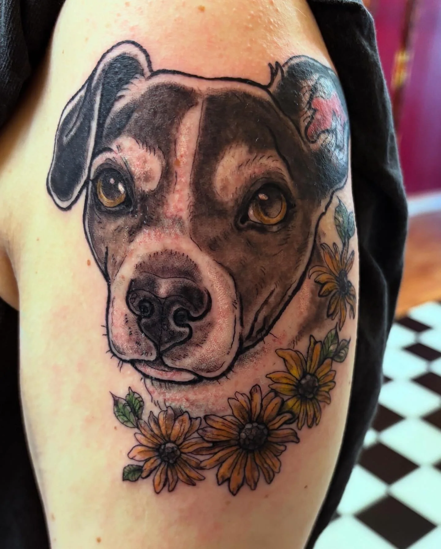Tattoo of a dog's face with detailed shading and yellow eyes, surrounded by sunflowers on the person's upper arm.