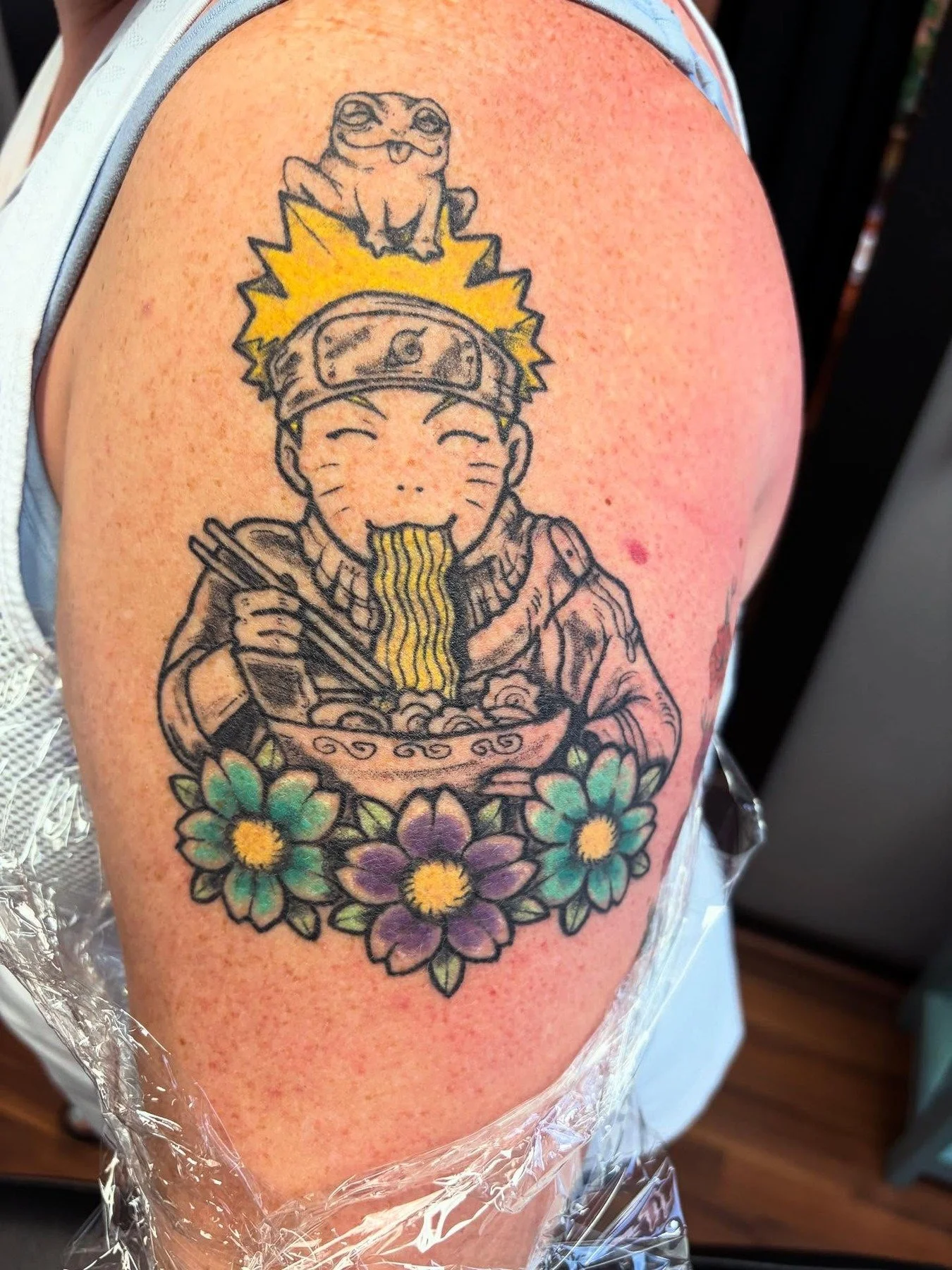 Colorful tattoo of a smiling anime-style character with whiskers, surrounded by flowers, holding chopsticks and a bowl of noodles, with a frog sitting on top of their head.