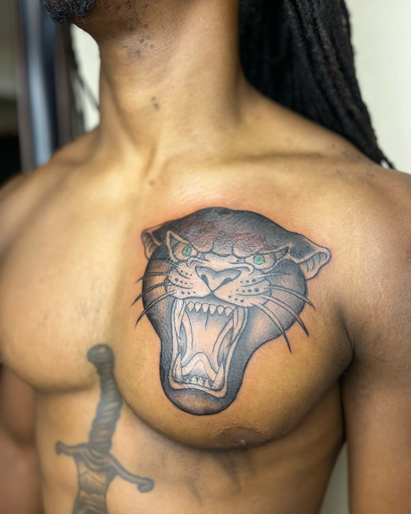 A close-up of a person's chest and neck showing a black and gray tattoo of a roaring panther head with green eyes, with additional tattoos visible on the shoulder and arm.