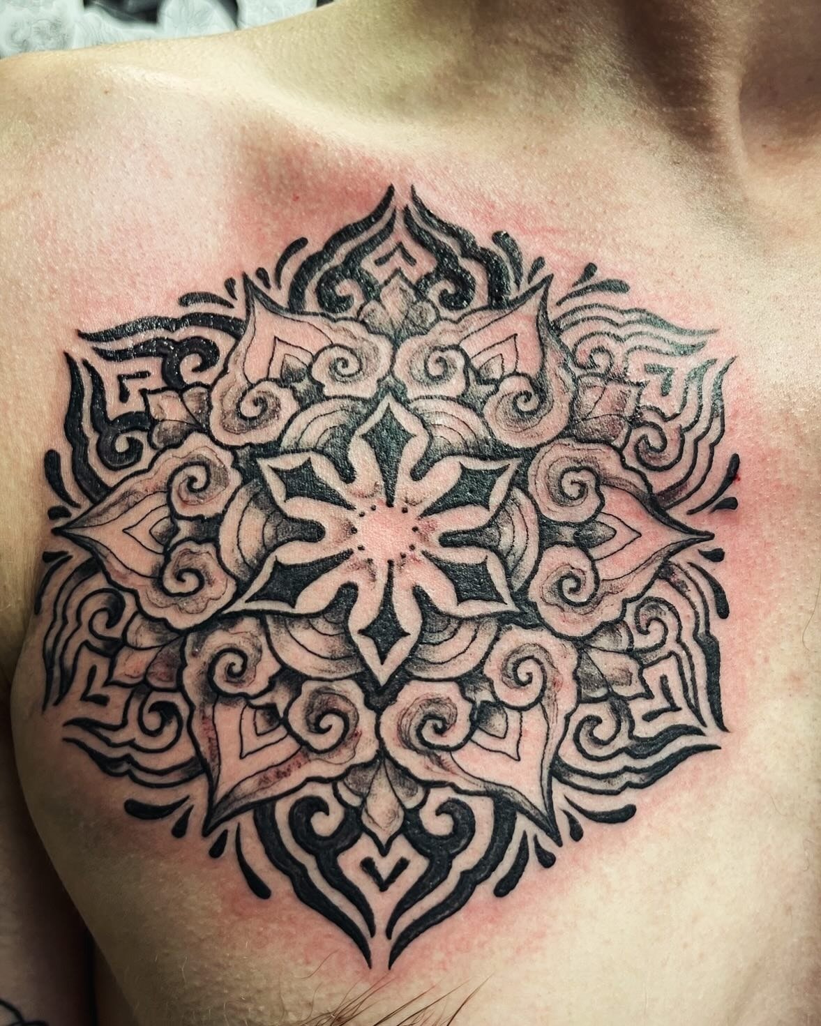 A detailed black and gray tattoo with a mandala-like design on a person's shoulder, featuring intricate geometric and swirling patterns.
