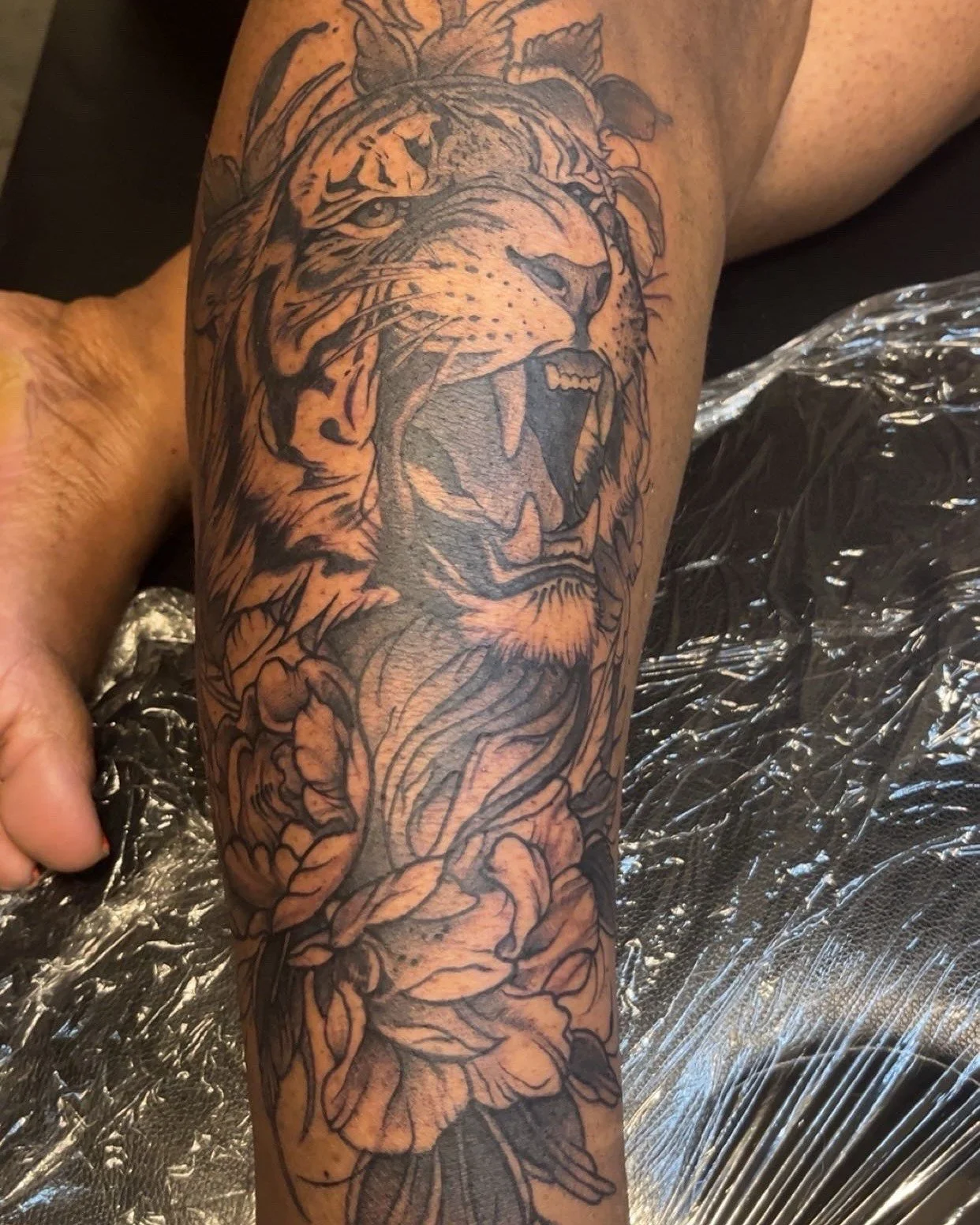 Black and gray tattoo of a roaring lion's head on a person's arm, surrounded by floral elements and leaves.