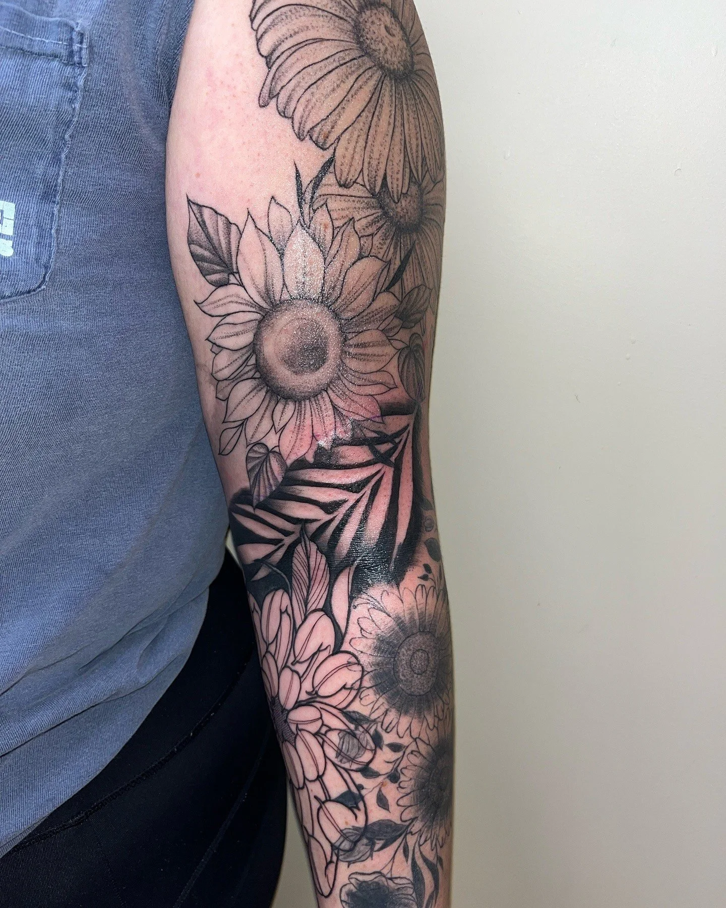 Black and gray floral tattoo sleeve on person's arm, featuring sunflowers and daisies, extending from shoulder to forearm, with detailed line work and shading.