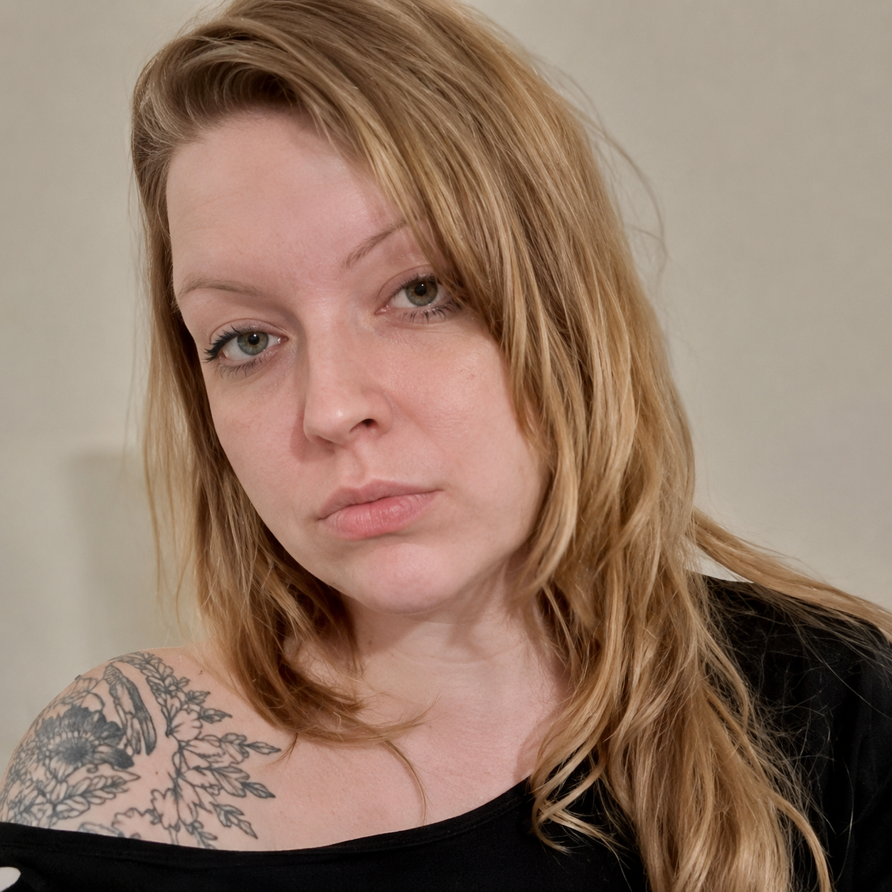 A woman with reddish-brown hair and light skin looking at the camera, wearing a black top with a tattoo on her left shoulder, against a plain beige background.