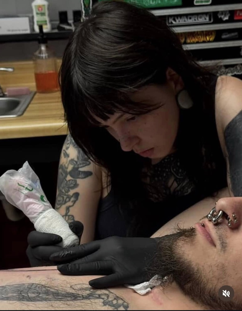 A woman giving a tattoo to a man on his chest. The woman has dark hair, tattoos on her arm, and is wearing black gloves. The man has facial hair, piercings on his nose, and tattoos on his chest.