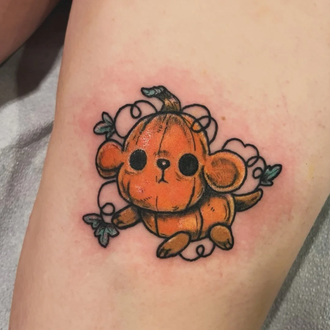 Tattoo of a cute pumpkin with a teddy bear face, surrounded by leaves and vines, on human skin.