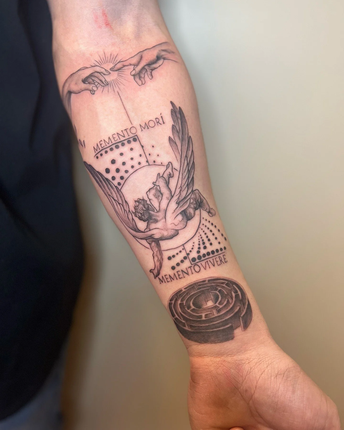 A black and gray tattoo on a person's arm depicting two hands reaching towards each other with radiating lines, a moon, wings, and Latin words 'Memento Mori' and 'Memento Vivere' along with geometric and dotted patterns.