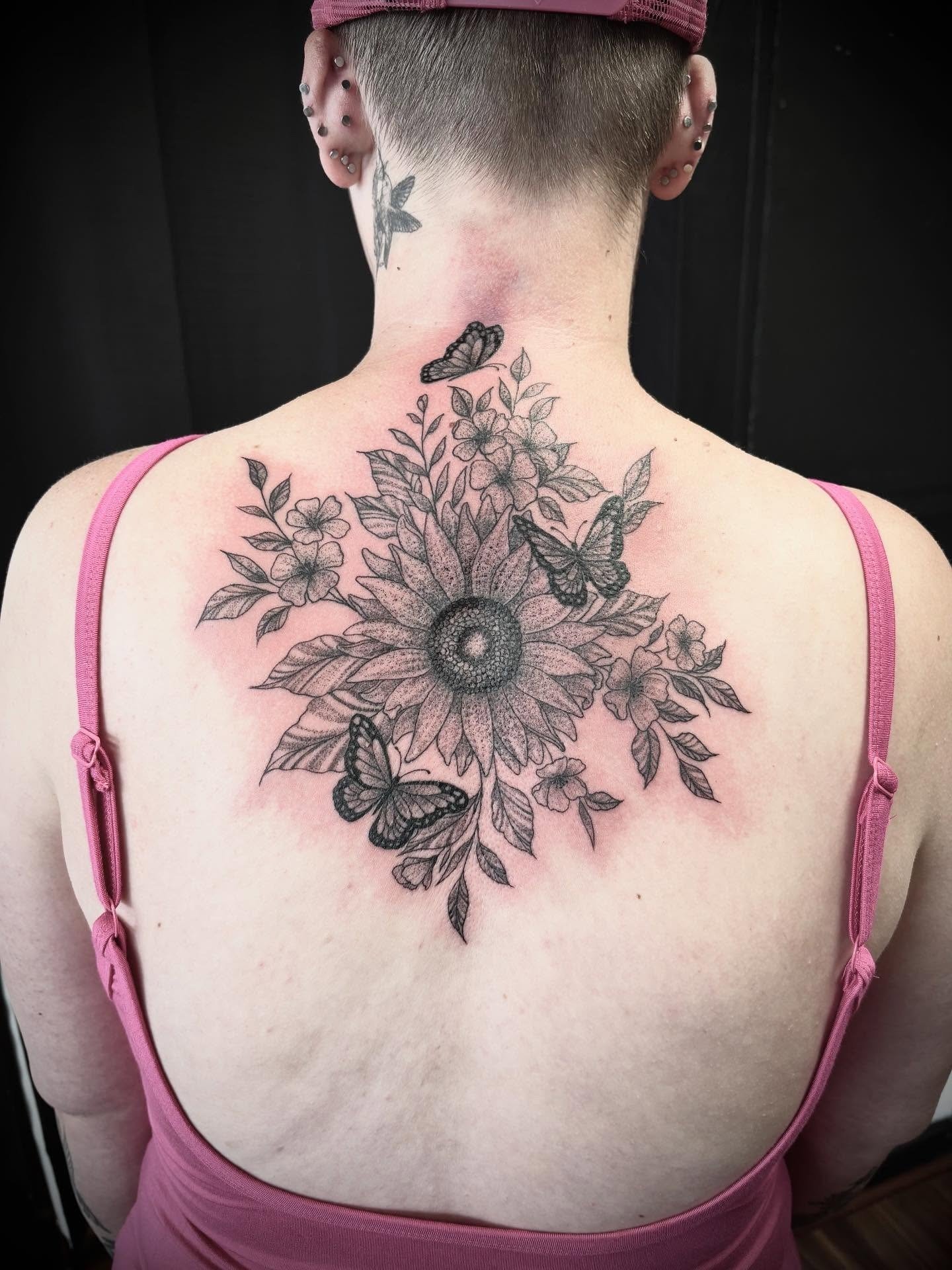 Back view of a woman with tattoos of flowers and butterflies on her upper back and neck, wearing a pink strappy top and a pink headband.