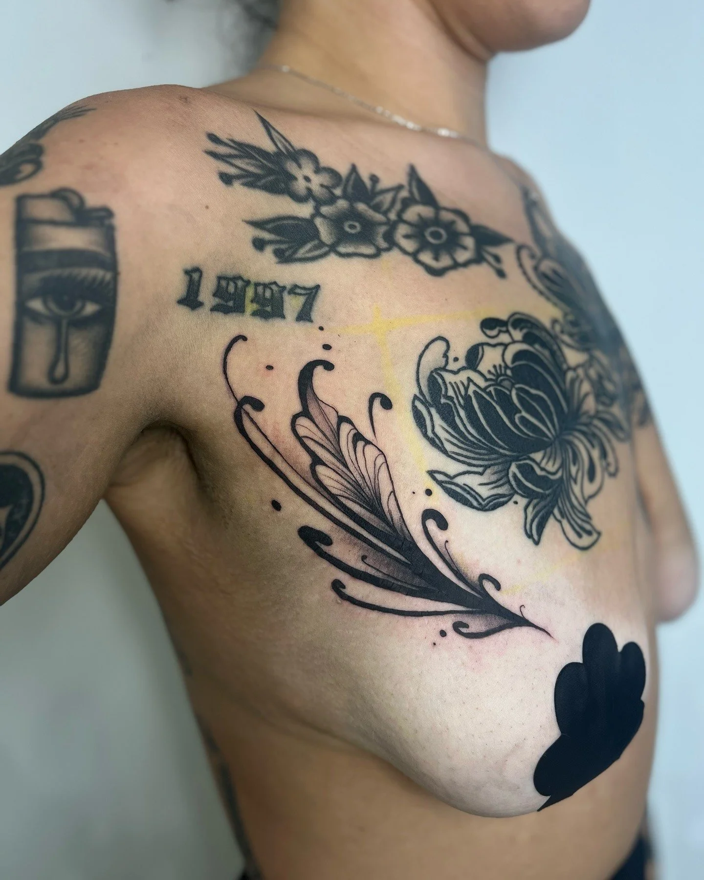 Close-up of a person's upper body with black tattoos, including flowers, the year 1997, an eye with teardrop, and other decorative designs.