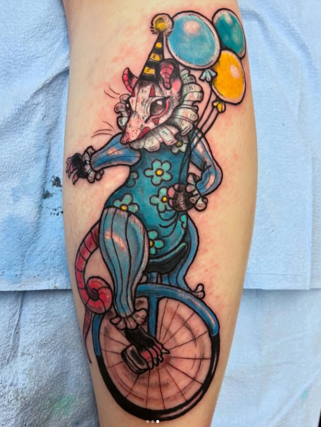 Colorful tattoo of a witch-like mouse riding a unicycle, wearing a blue dress with yellow flowers, holding balloons, with a striped party hat and a ruffled collar.