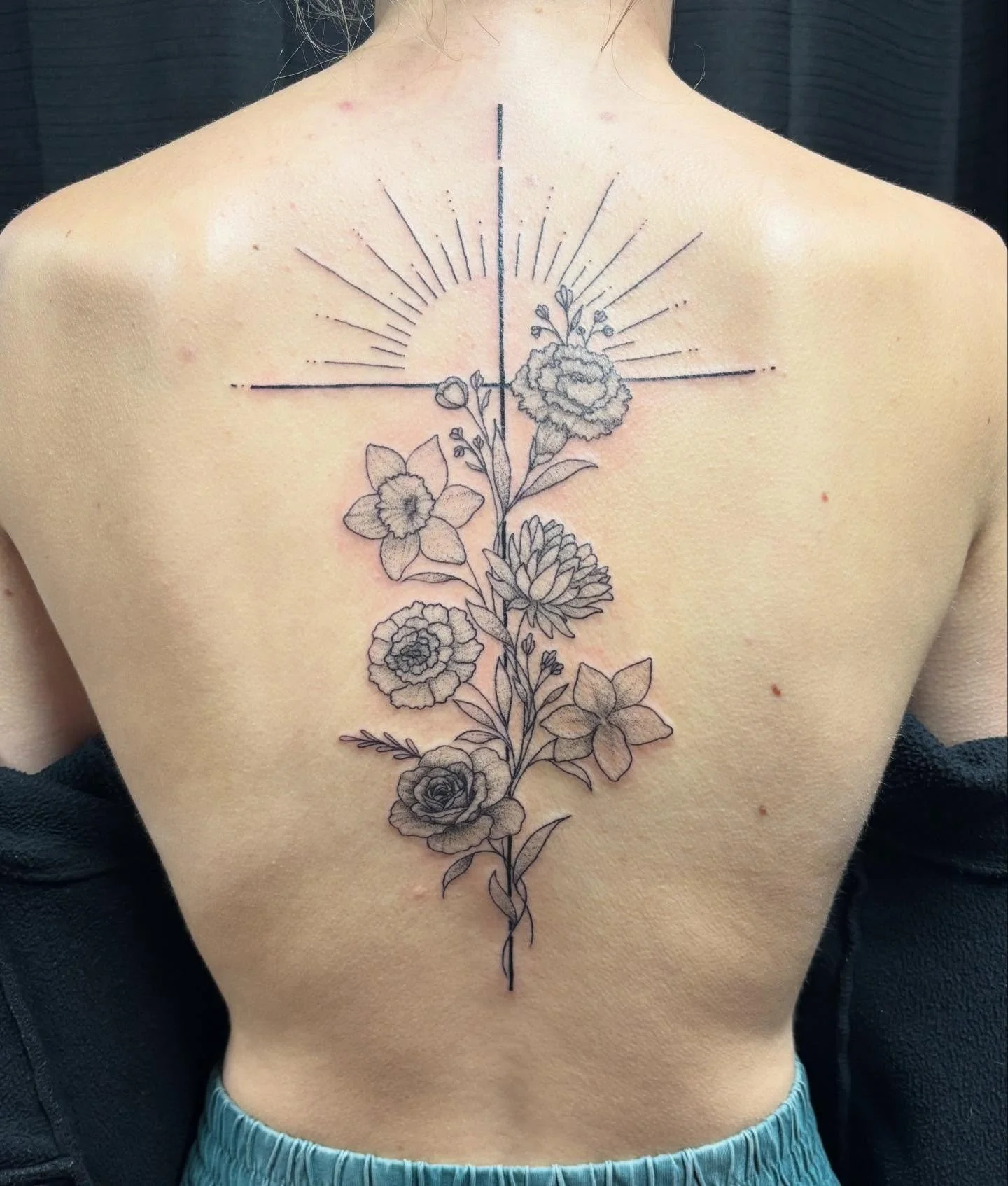A person's back with a tattoo design featuring a vertical and horizontal line, a small sunburst, and a detailed floral arrangement with roses and other flowers.