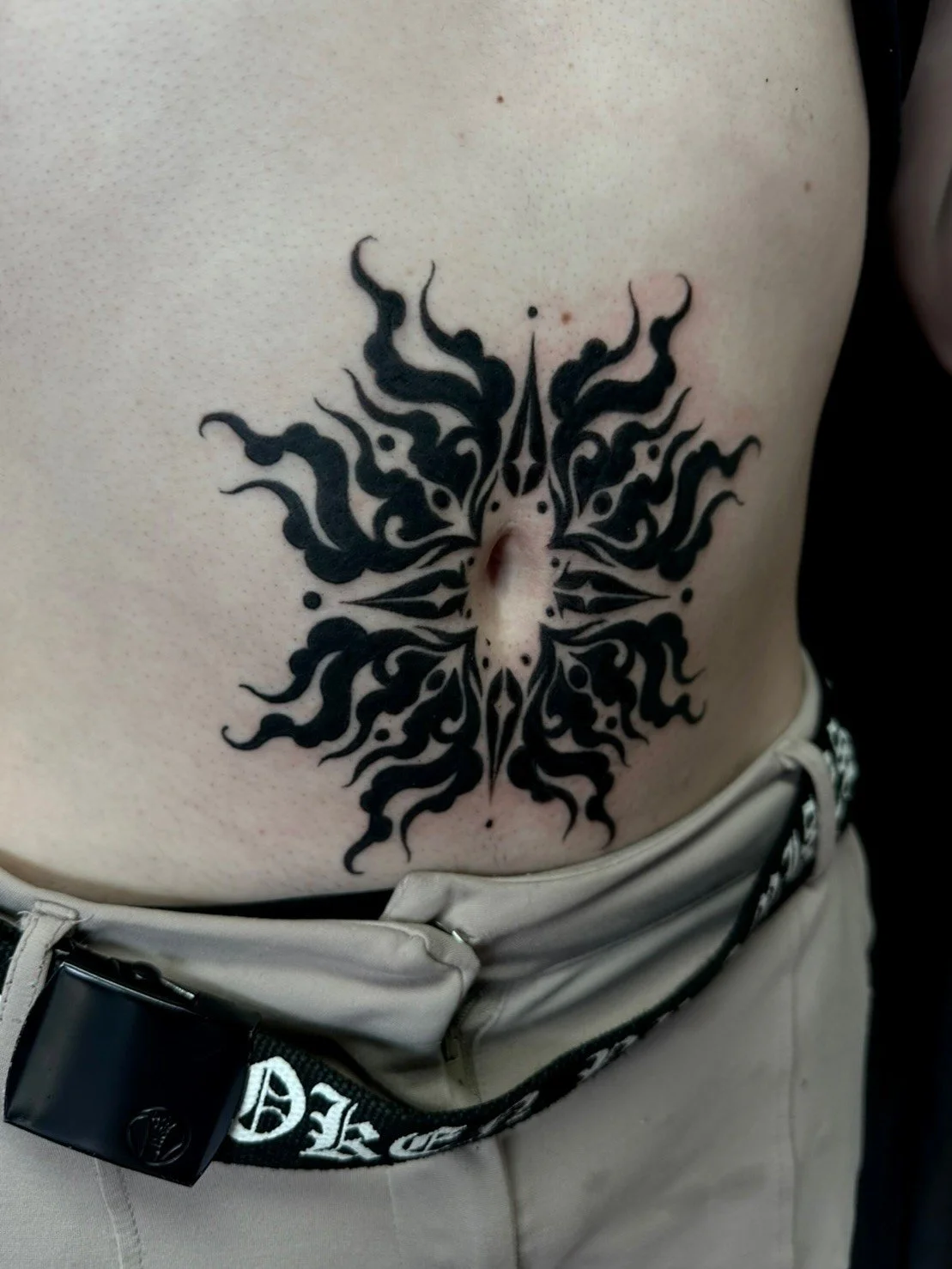 A black tribal-style tattoo on a person's lower abdomen, featuring symmetrical, flame-like shapes aligned around the belly button.