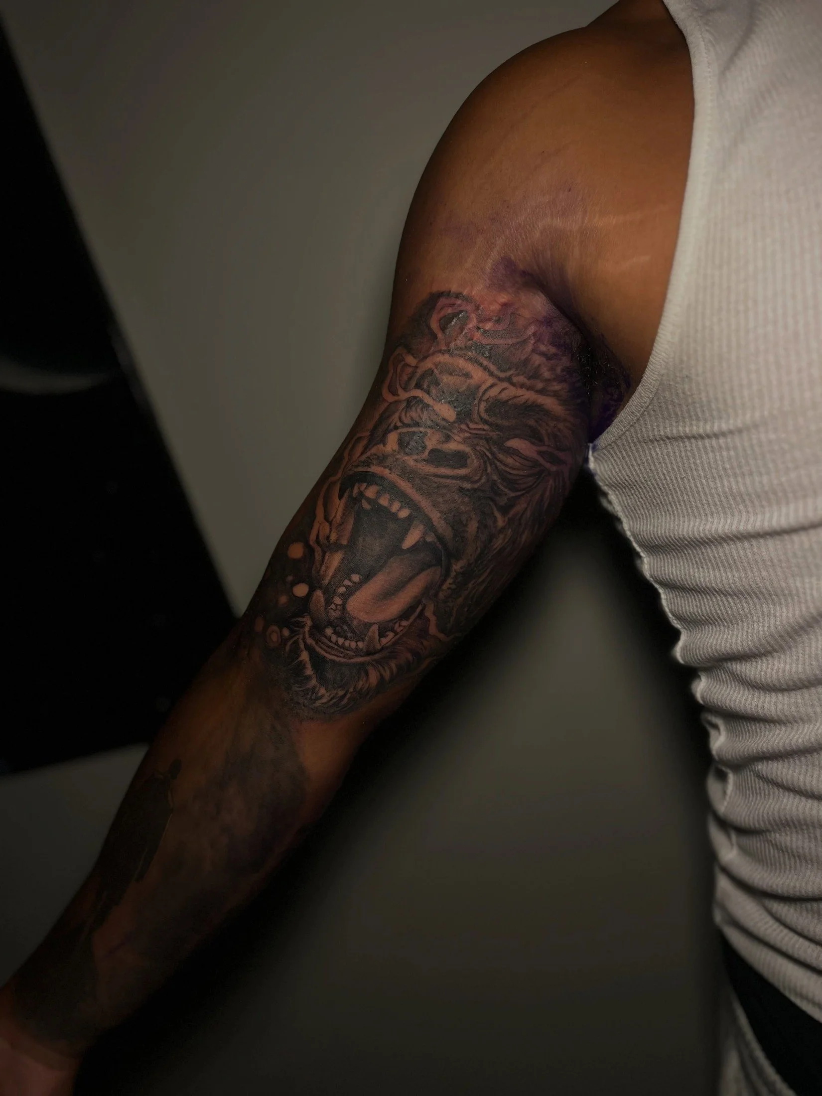 A close-up of a person's tattooed arm resting on a surface, with visible shoulder and white sleeveless shirt.