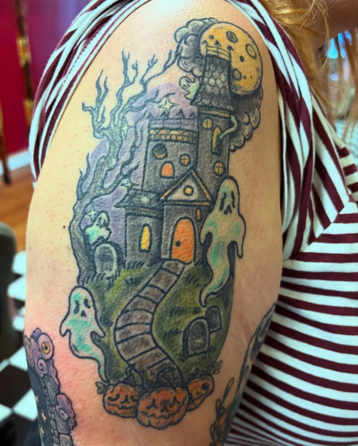 Tattoo of a haunted house with spooky trees, a winding staircase, clouds, and ghosts on upper arm of a person wearing a striped shirt.