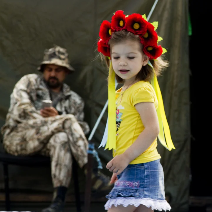 A young girl wearing a Vinok, a wreath of poppies that is part of traditional Ukrainian folk costume, explores the wreckage of the barricades and tent city on the Maidan in central Kyiv, while a dissenter in camouflage relaxes outside his tent.