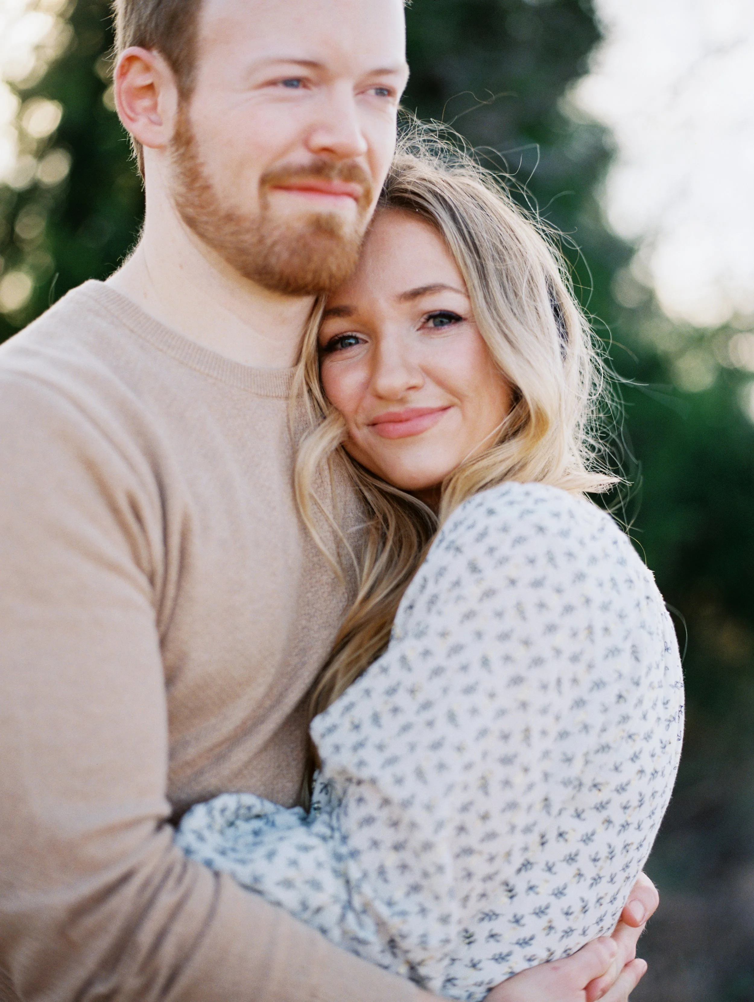 Close-up of a man and woman hugging outdoors. The man has a beard and is wearing a beige sweater, and the woman has wavy blonde hair and is wearing a white top with small blue pattern. They are smiling softly with a blurred background of greenery.