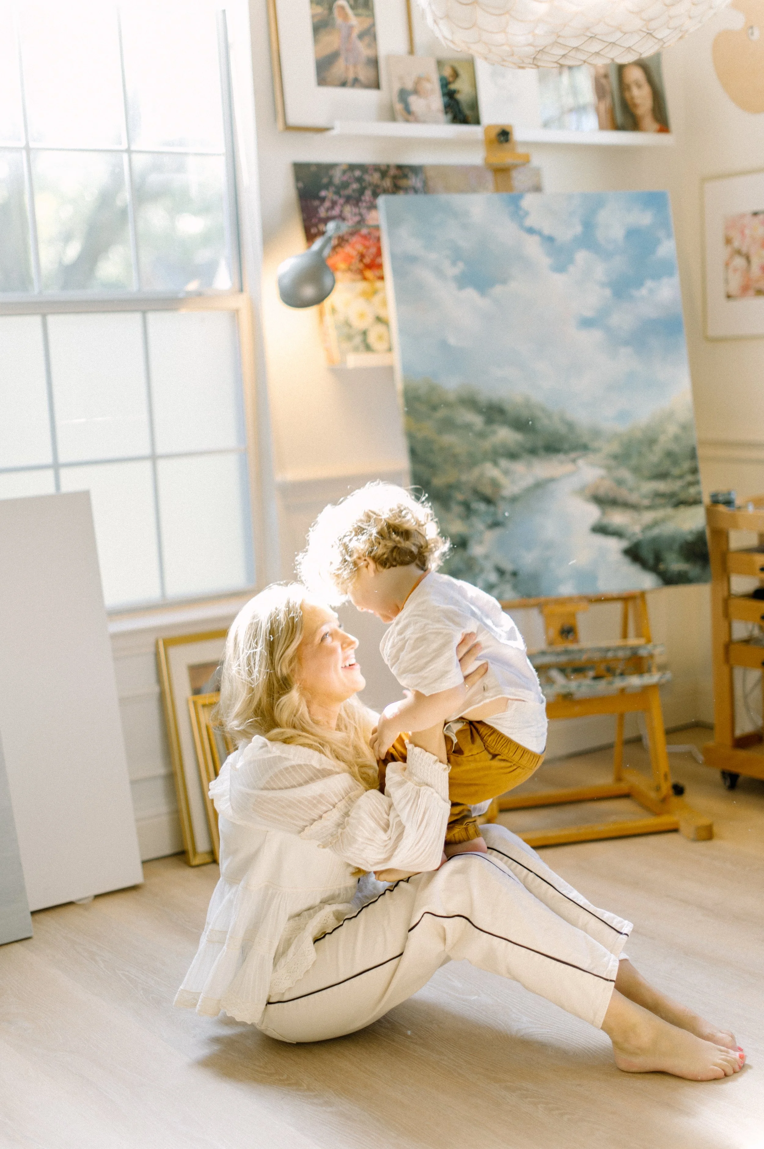 A woman with long blonde hair sitting on the floor and lifting a young boy with curly blonde hair, who is smiling and looking at her, in an art studio with paintings and large window in the background.