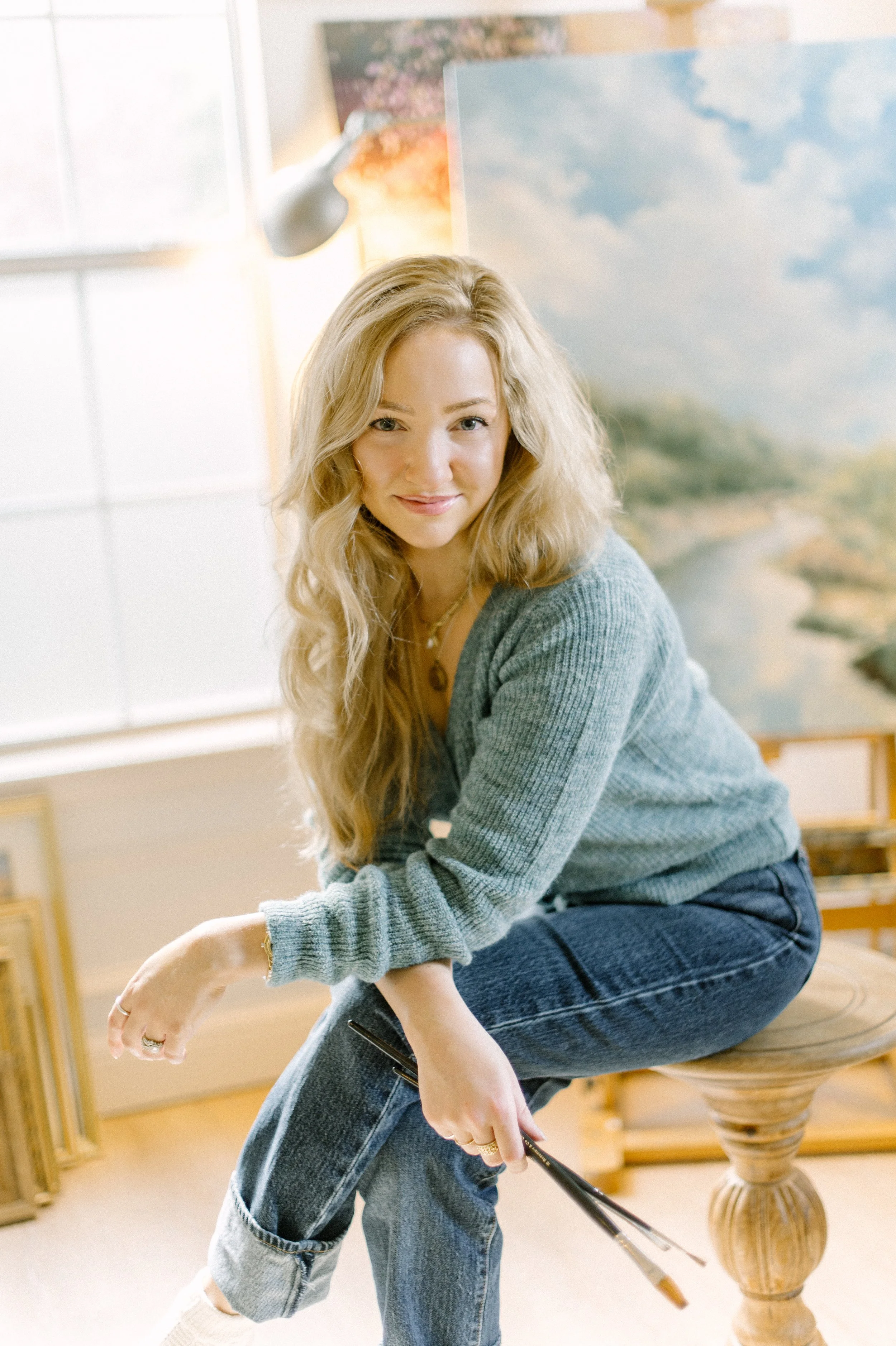 A woman with long blonde hair, wearing a gray sweater and blue jeans, sitting on a wooden stool with one leg crossed over the other. She is holding a paintbrush, smiling, and appears to be in a studio or art space with a landscape painting and window in the background.