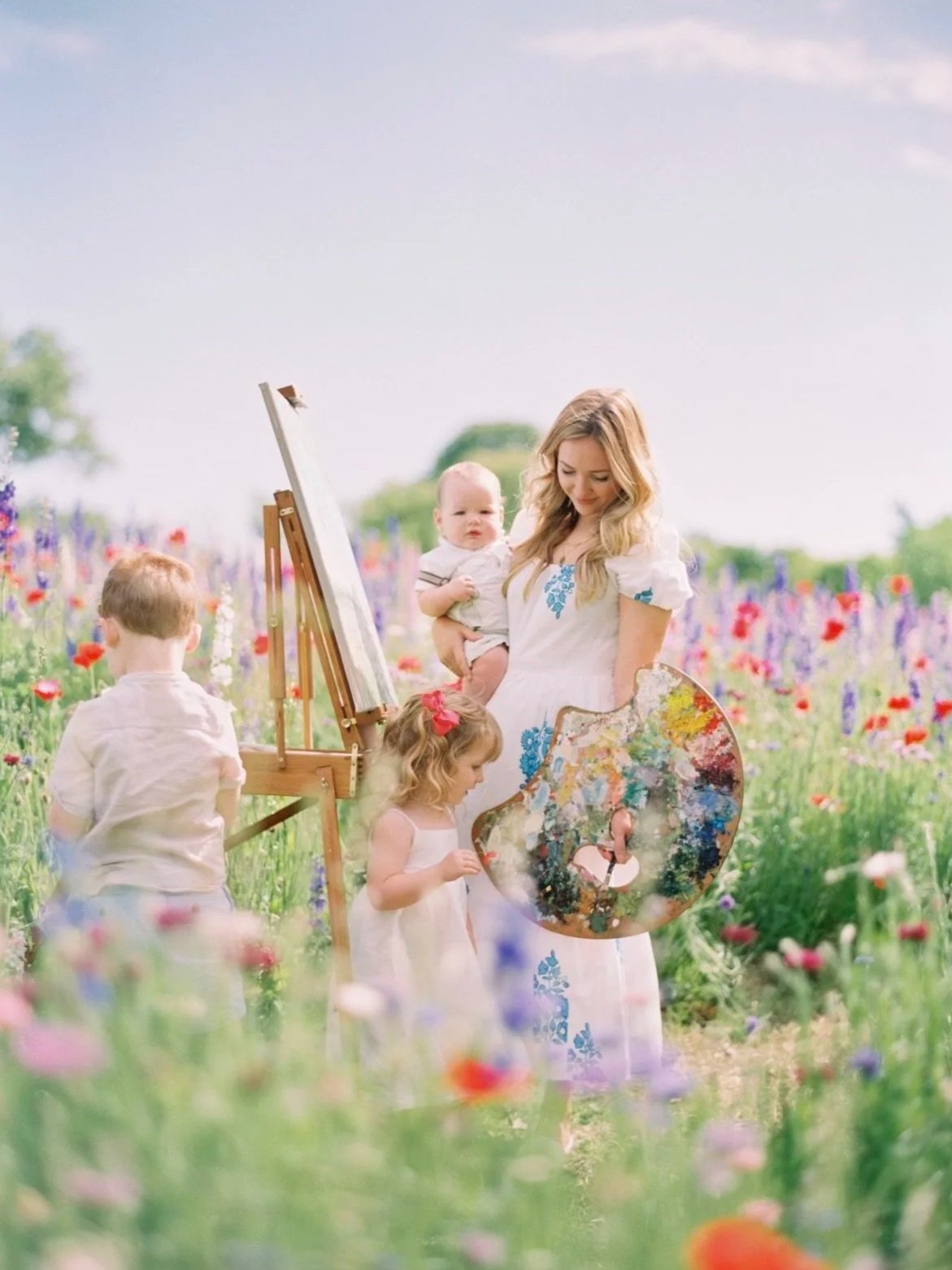 A woman and three children in a field of colorful flowers, with the woman holding a painter's palette and the children engaging around her, some near an easel with a blank canvas.