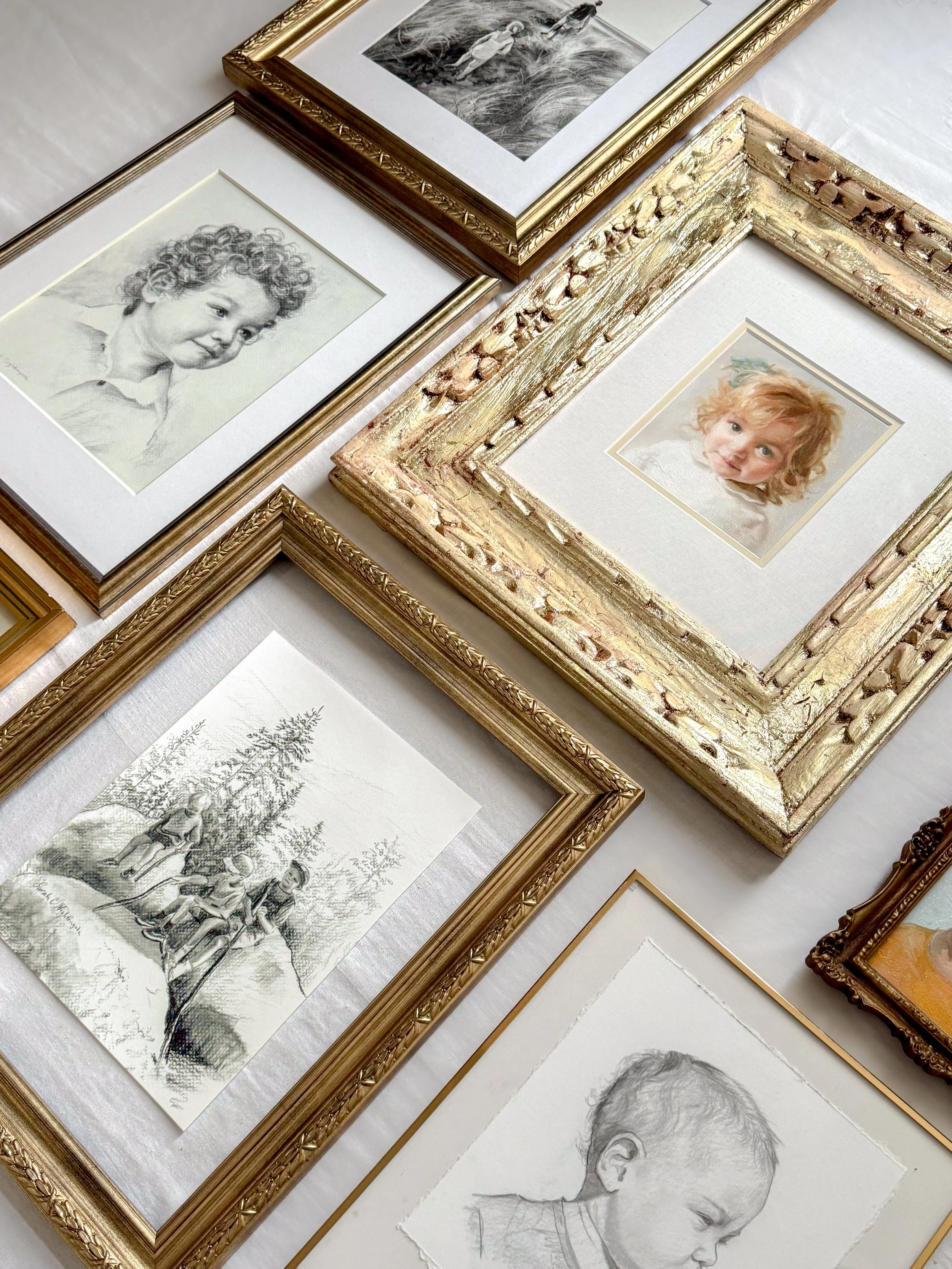 A collection of framed drawings and photographs arranged on a white surface, including a portrait of a young child with curly hair and a black-and-white illustration of children playing outdoors.