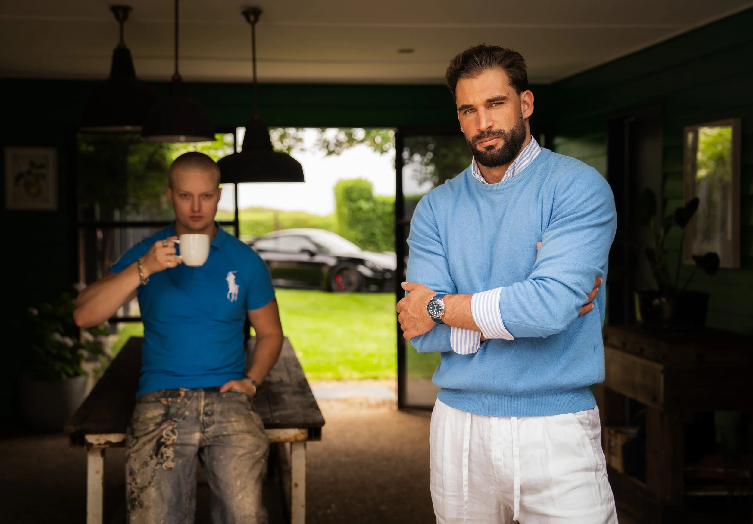 Two men in a room with large open doors and a garden view in the background. The man on the right, with dark hair and beard, is standing with arms crossed, wearing a light blue sweater, striped shirt, white pants, and a wristwatch. The man on the lef