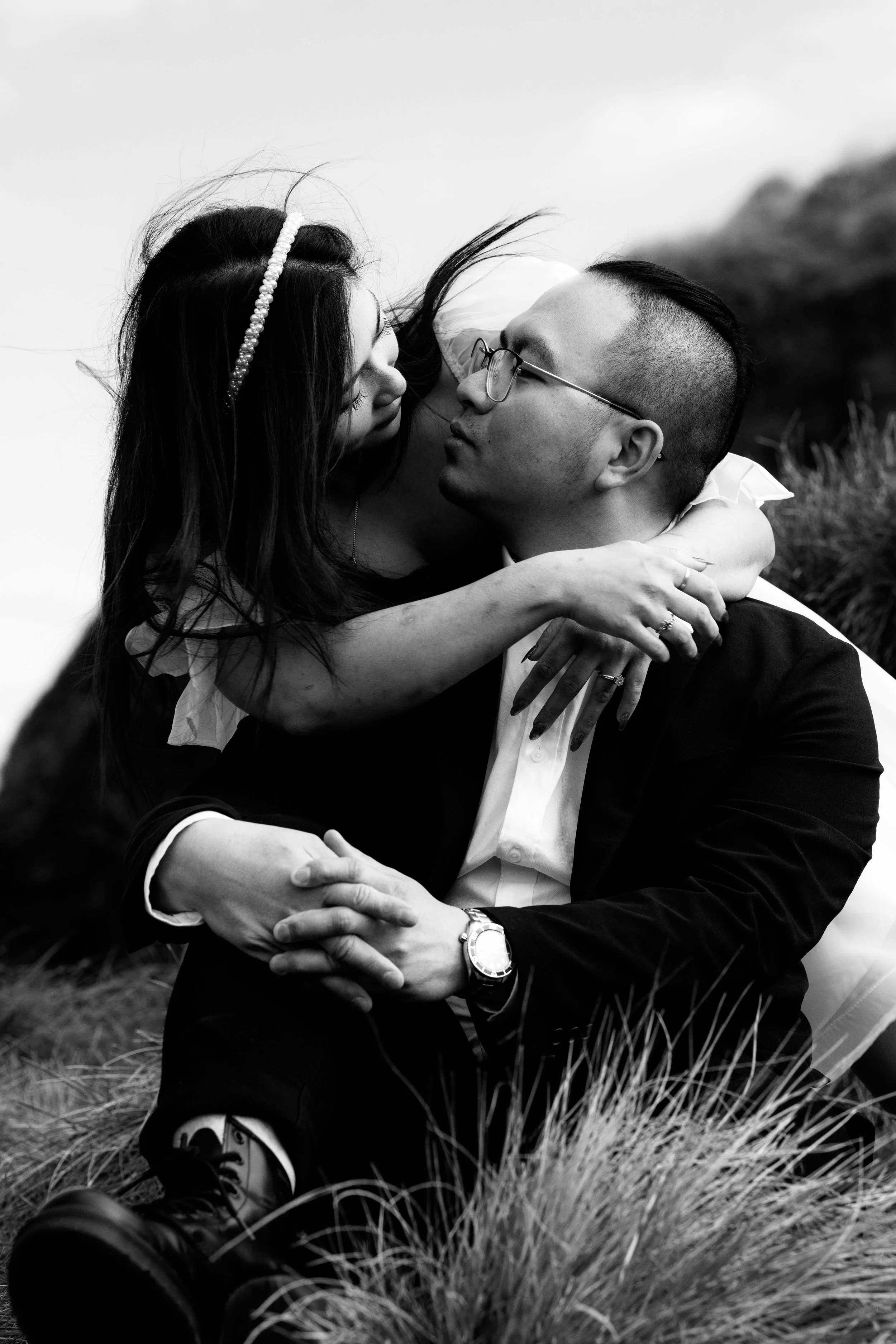 A black and white photo of a couple embracing outdoors, with the woman leaning in to kiss the man on the face, who is sitting on the grass and wearing a suit and glasses.