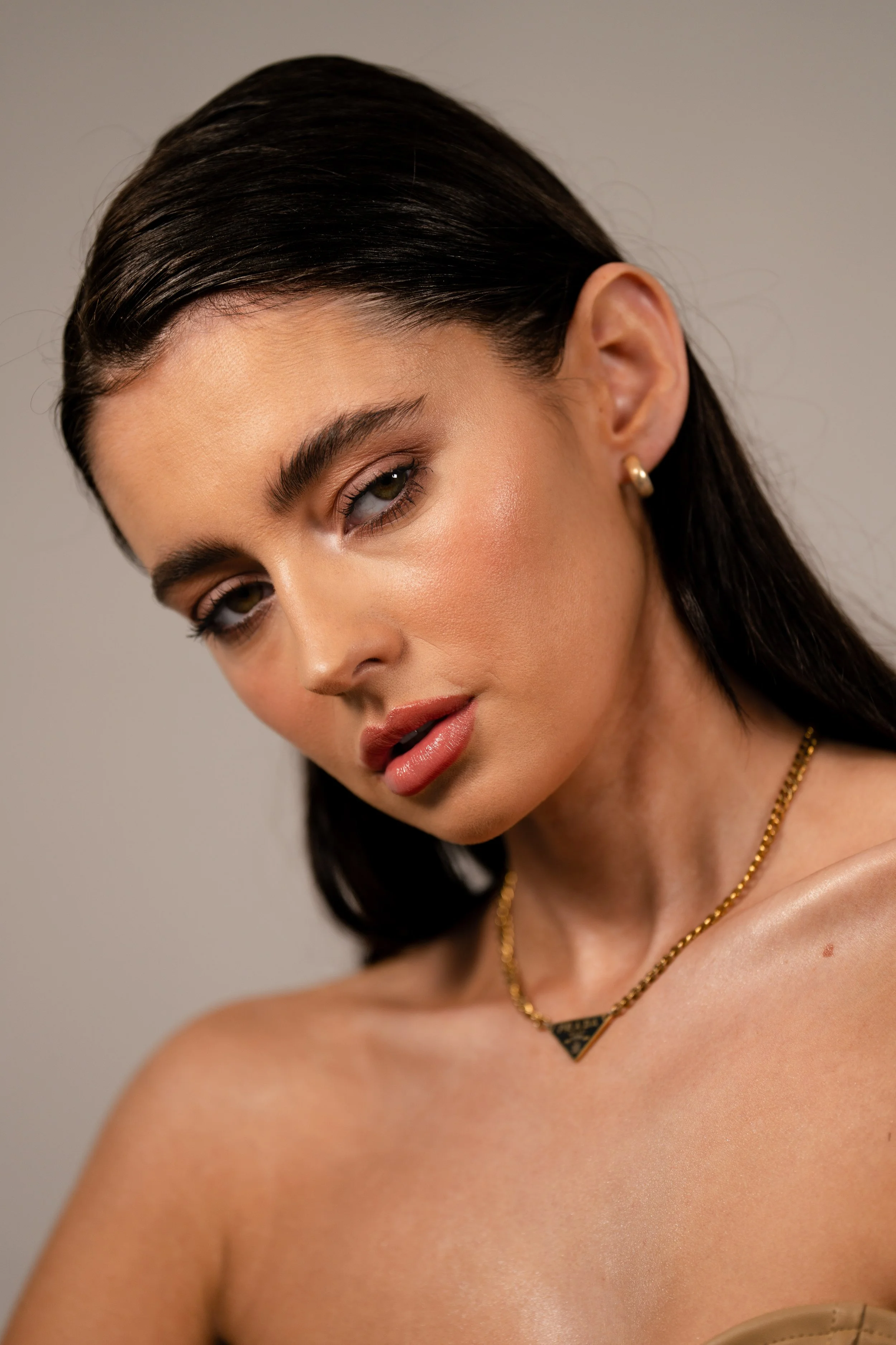 A close-up of a woman with smooth, dark hair, wearing gold earrings and a necklace, with makeup emphasizing her eyes and lips, against a neutral background.