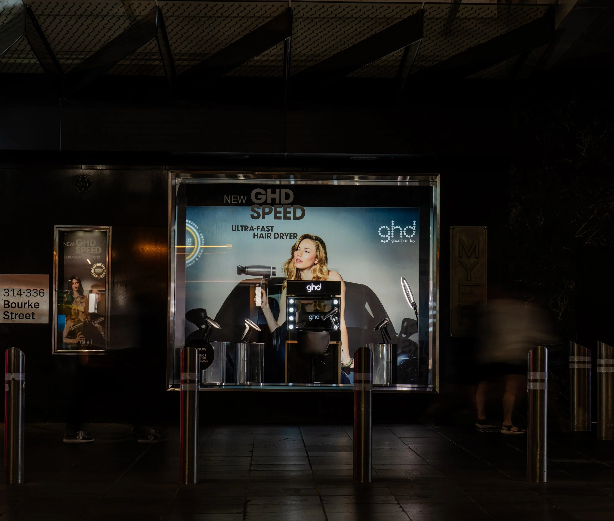 Advertisement display for GHD Speed ultra-fast hair dryer featuring a woman holding a hairdryer in front of a promotional backdrop, with other hair styling tools around her.