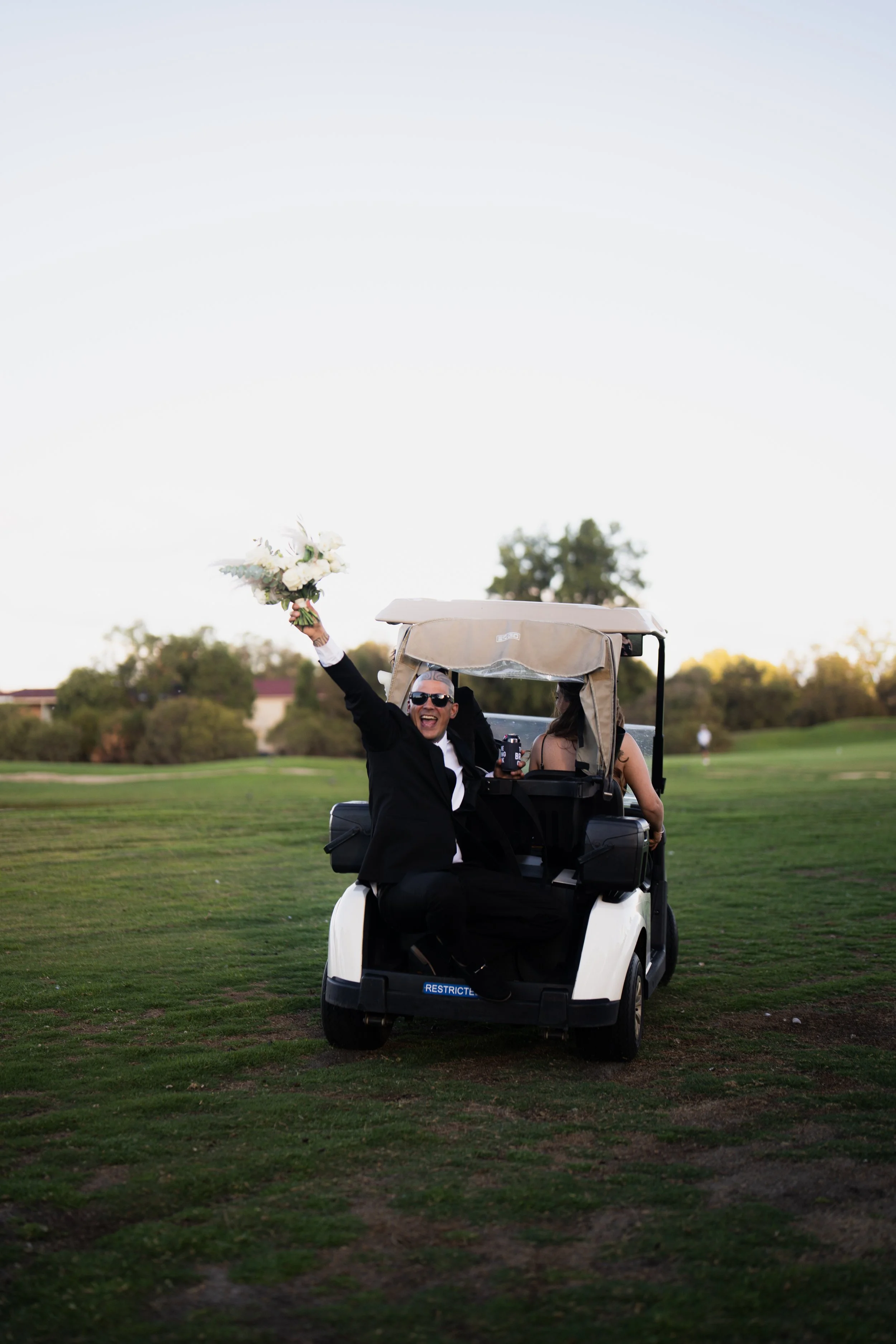 A man in a tuxedo and sunglasses sitting in a golf cart on a golf course, holding a bouquet of white flowers and smiling happily.