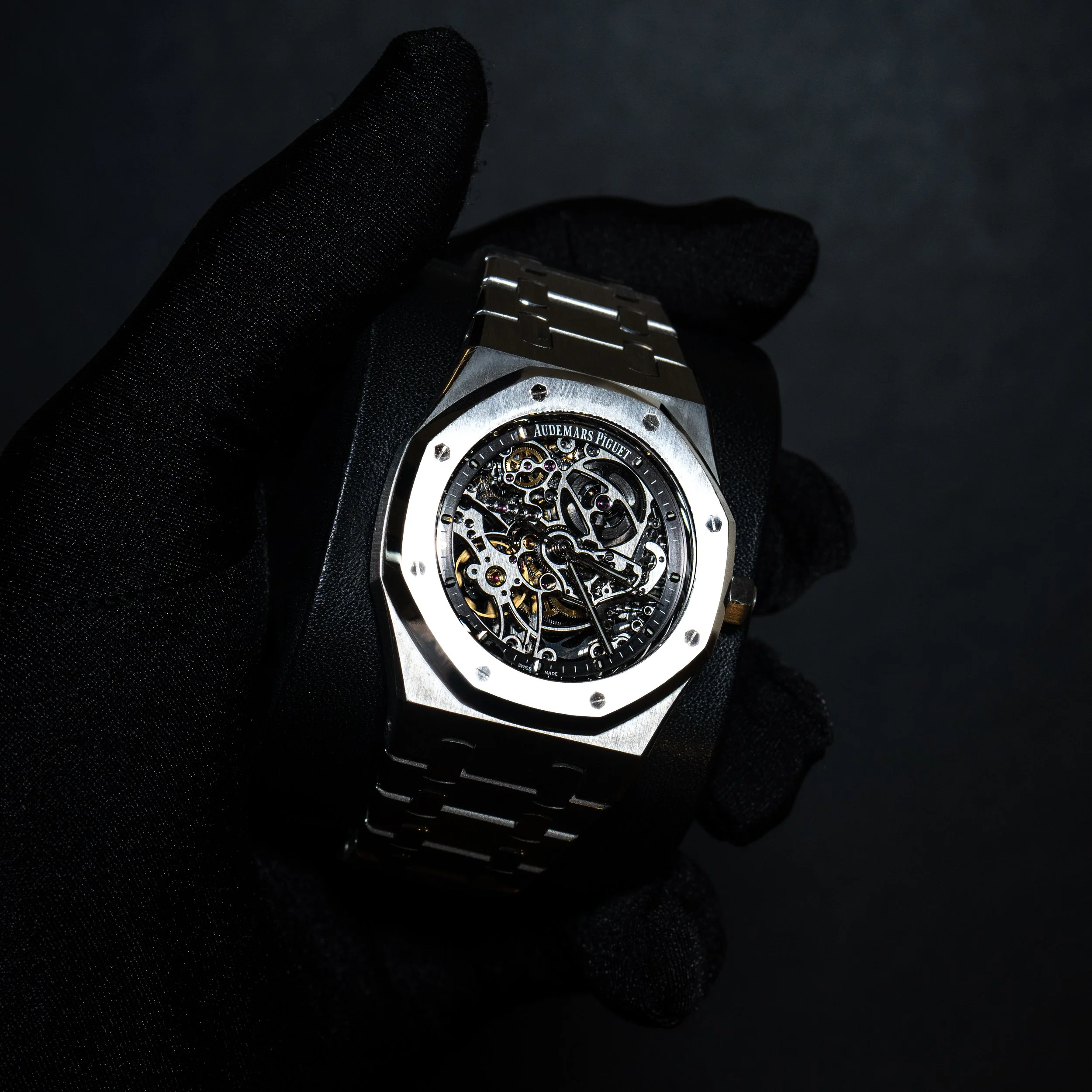 A luxury skeleton wristwatch with a metal band, held by a gloved hand against a dark background.
