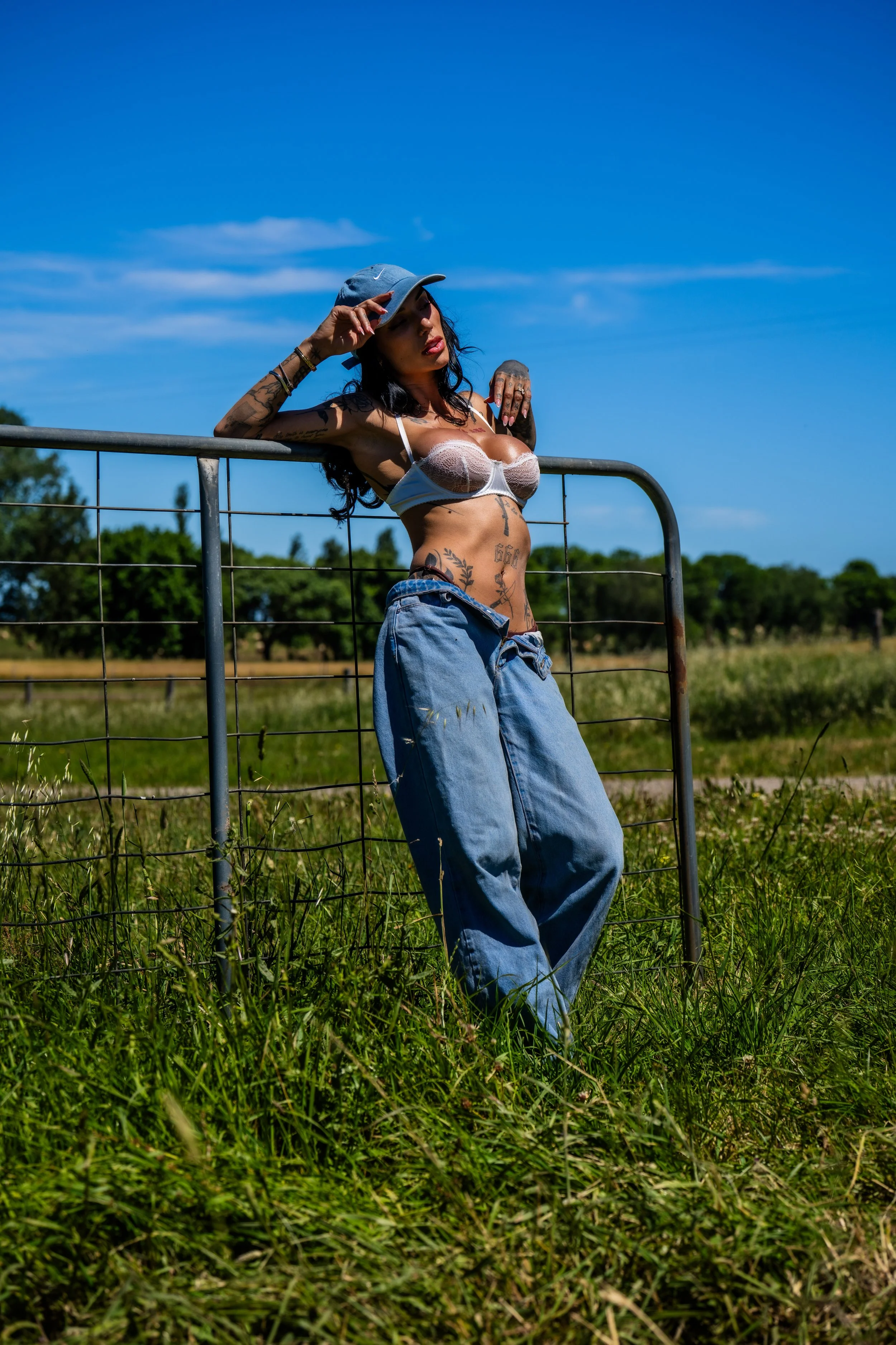A woman standing outdoors in a grassy field, leaning against a metal fence, wearing a cap, a sheer white bra, and loose denim pants, with tattoos on her torso and arms, under a bright blue sky.