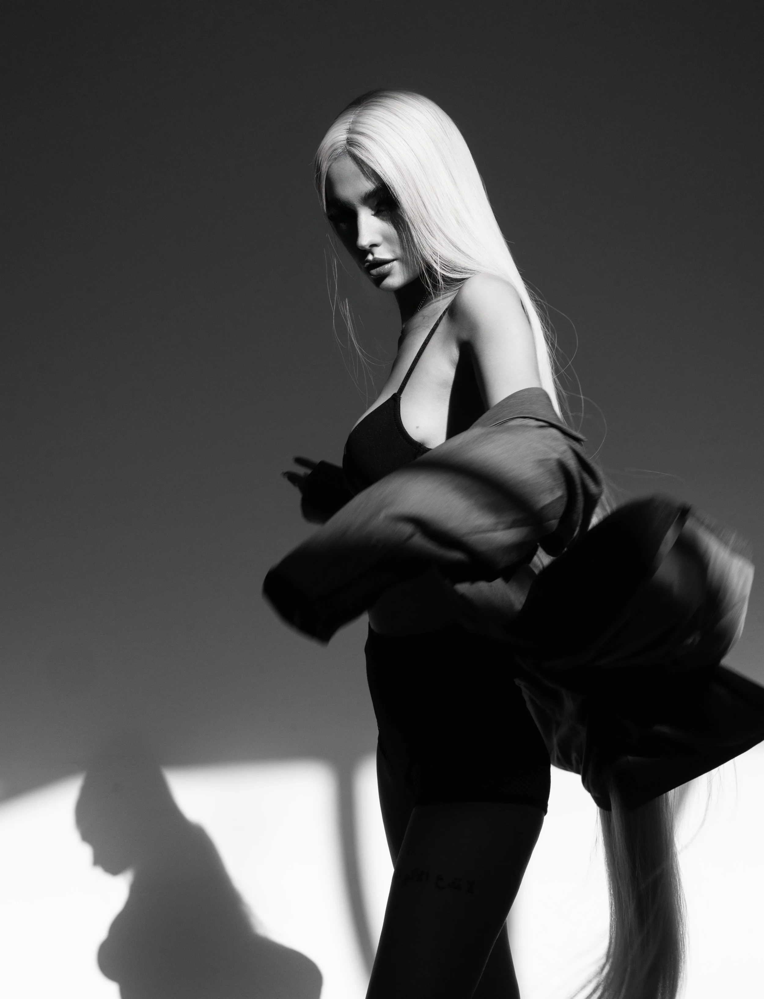 Black and white photo of a woman with long blonde hair, wearing a black top and shorts, with a jacket draped over her shoulders, posing against a plain background with shadow play.