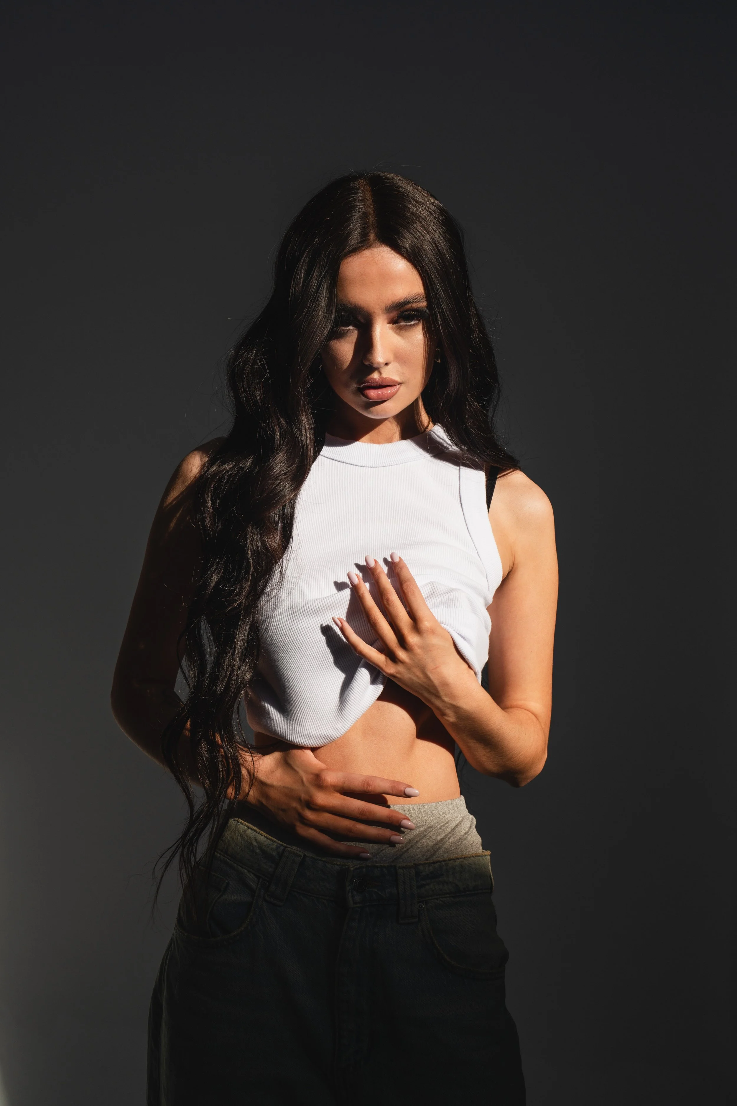 A woman with long dark hair wearing a white crop top and dark pants, standing against a dark background, with one hand on her chest and the other on her waist.