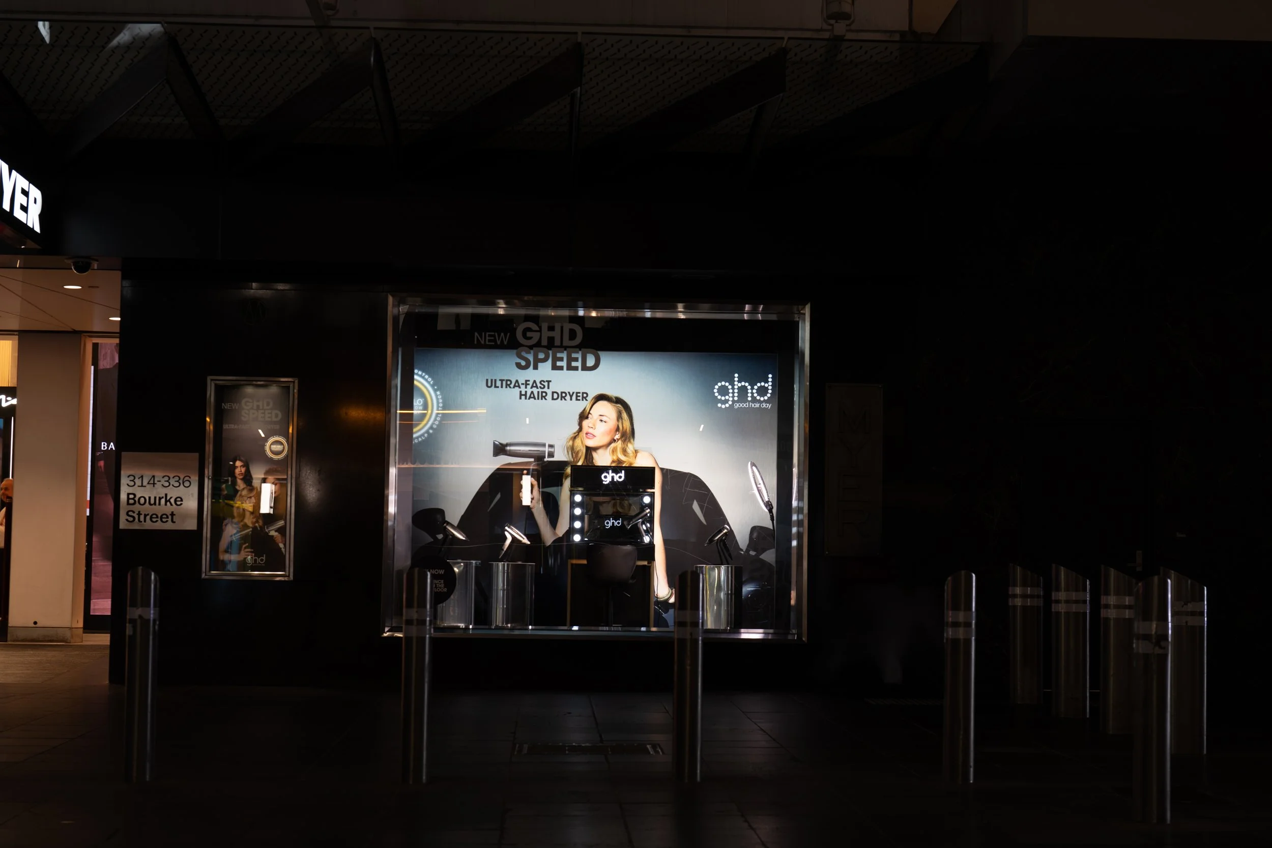 Nighttime store window display advertising GHD hair dryers, featuring a woman using a hair dryer, with promotional text and branding.