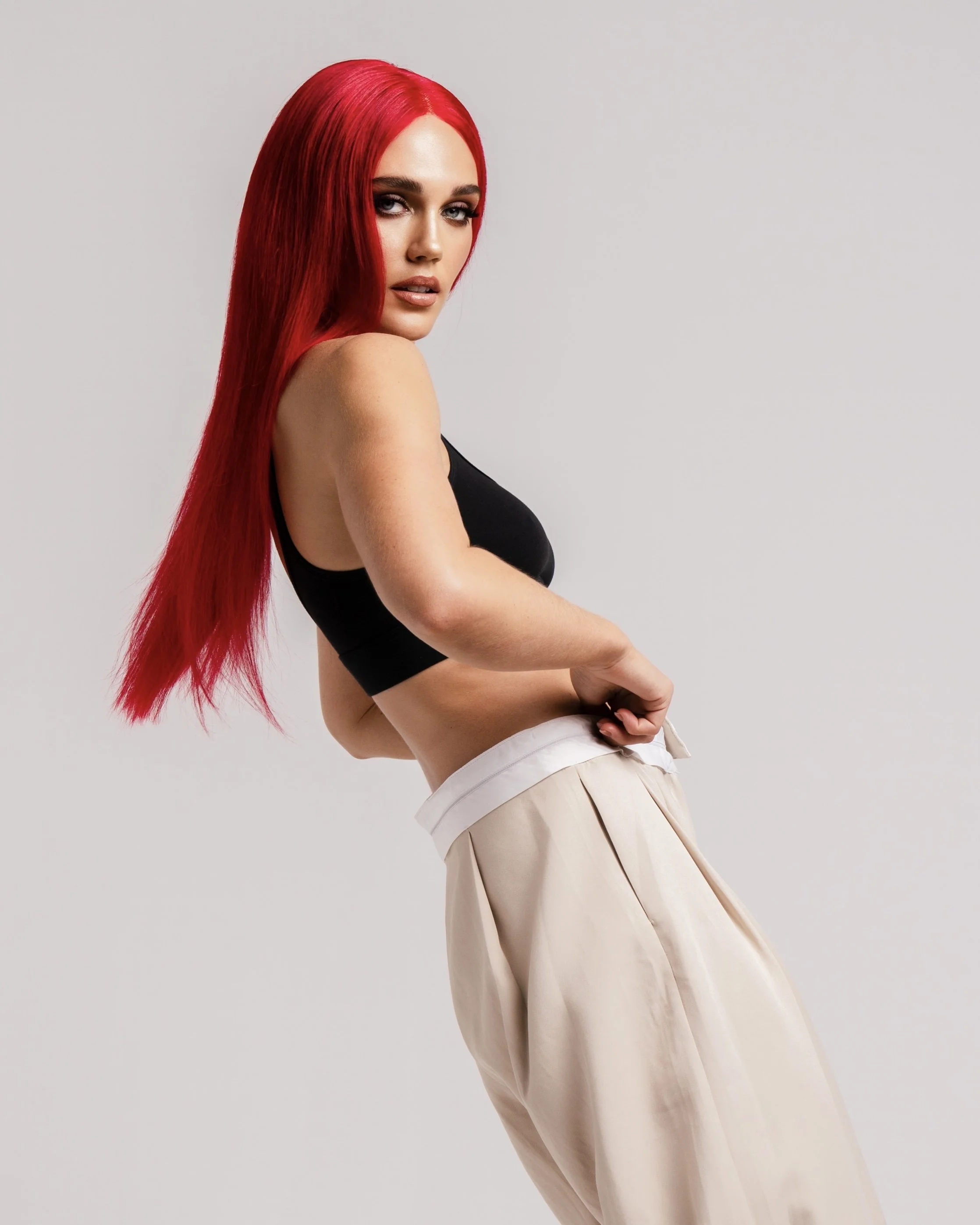 A woman with long red hair wearing a black sleeveless top and beige pants, standing against a plain background.