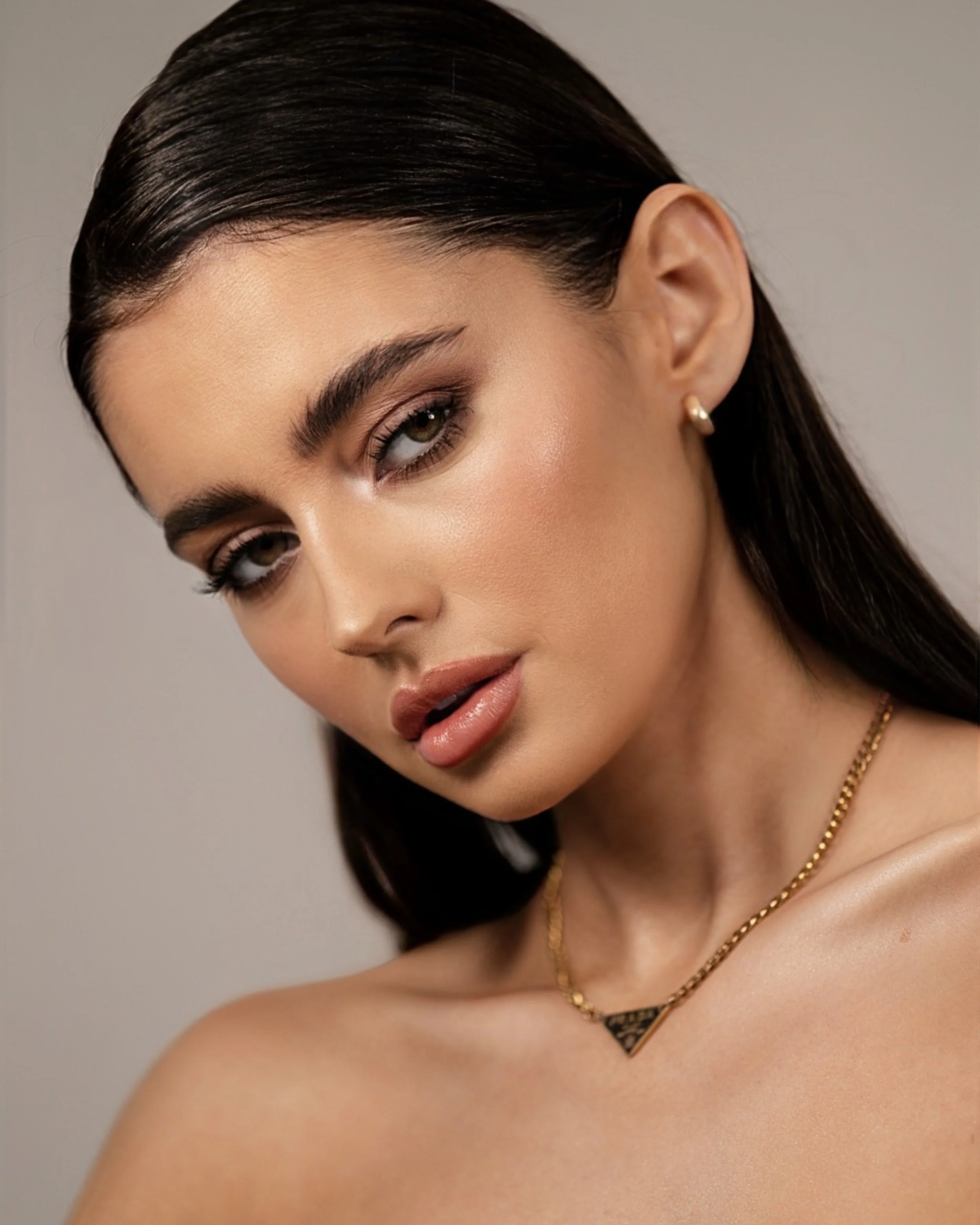 Close-up of a young woman with dark hair, wearing minimal makeup and a gold necklace, with a neutral background.