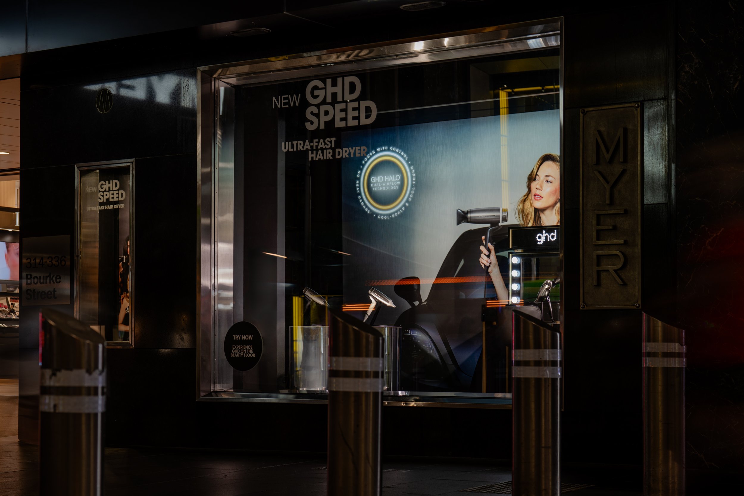 A store display showcasing the GHD Speed ultra-fast hair dryer with a woman holding the hair dryer in a digital advertisement behind glass.